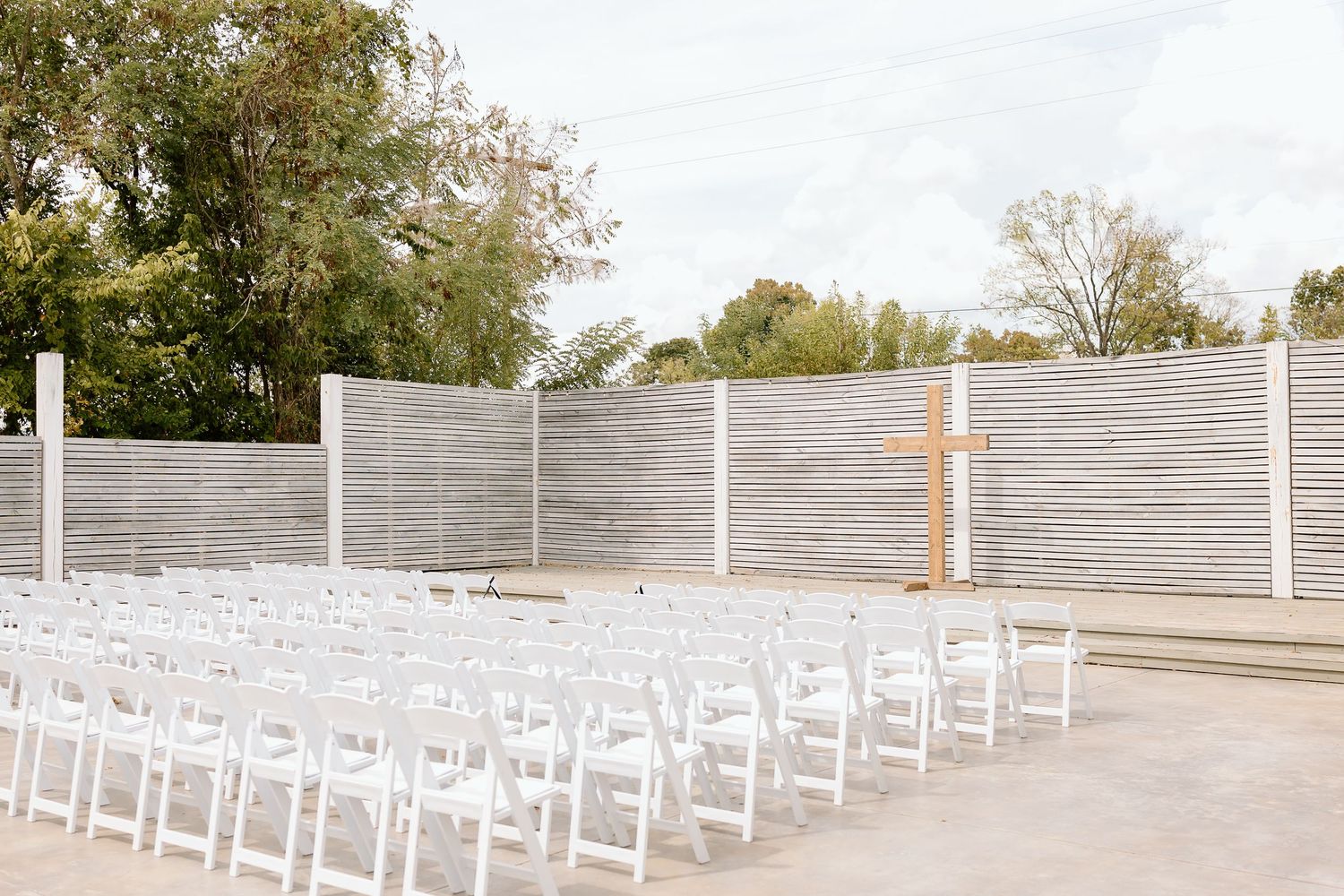 White wooden chairs arranged in rows for an outdoor ceremony on a concrete patio with trees in the background.