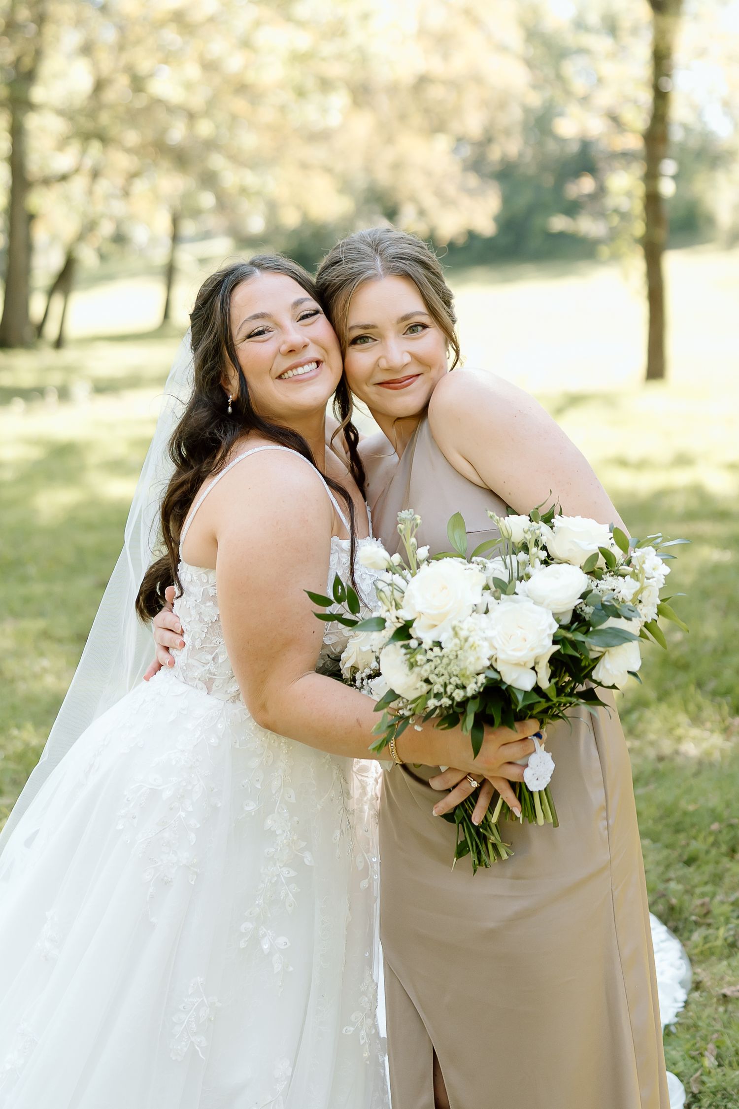 A bride in a white lace gown stands with a bridesmaid in beige holding a white floral bouquet outdoors.