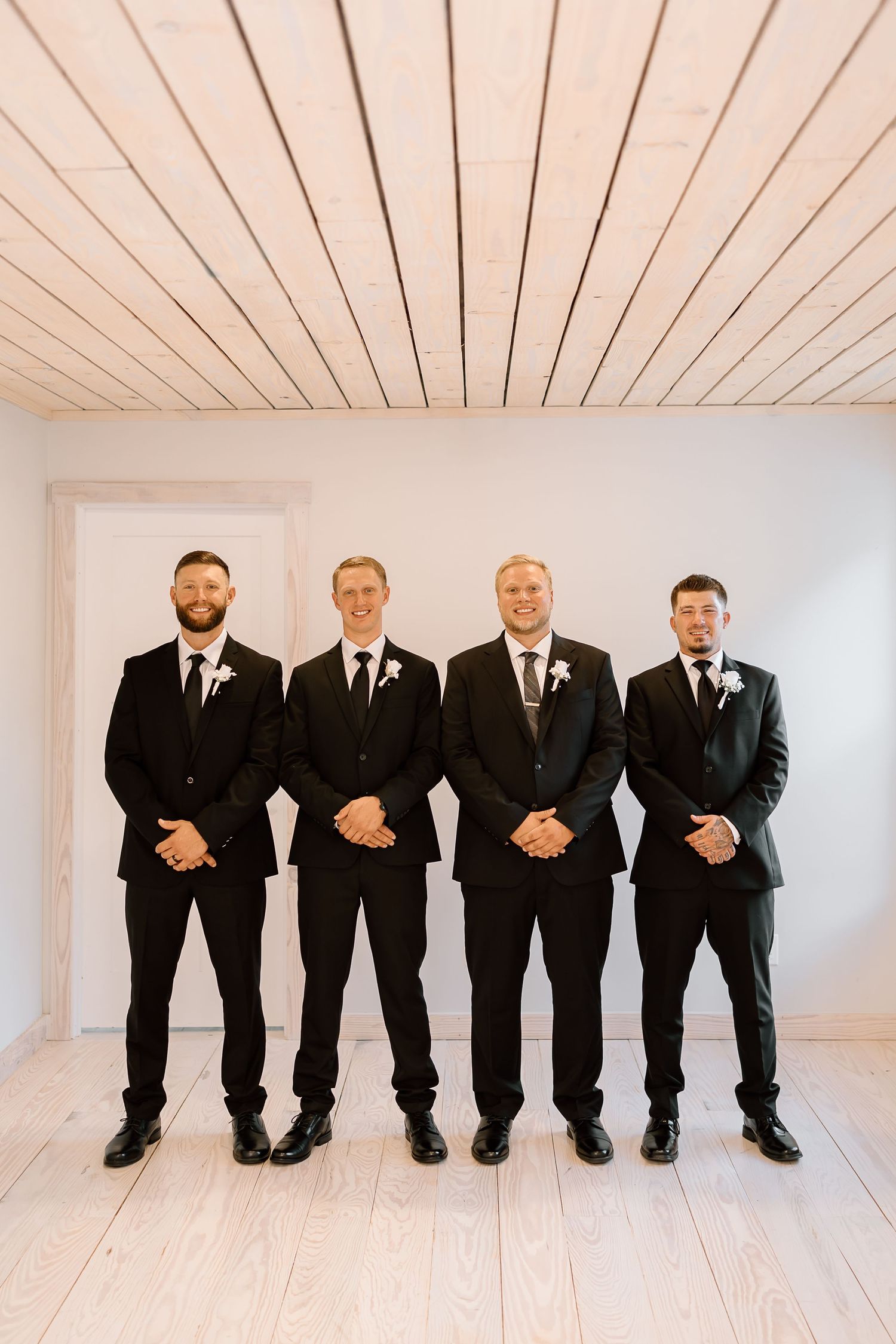 Four groomsmen in matching black suits and boutonnieres stand in a row against a white wall with wood ceiling details.