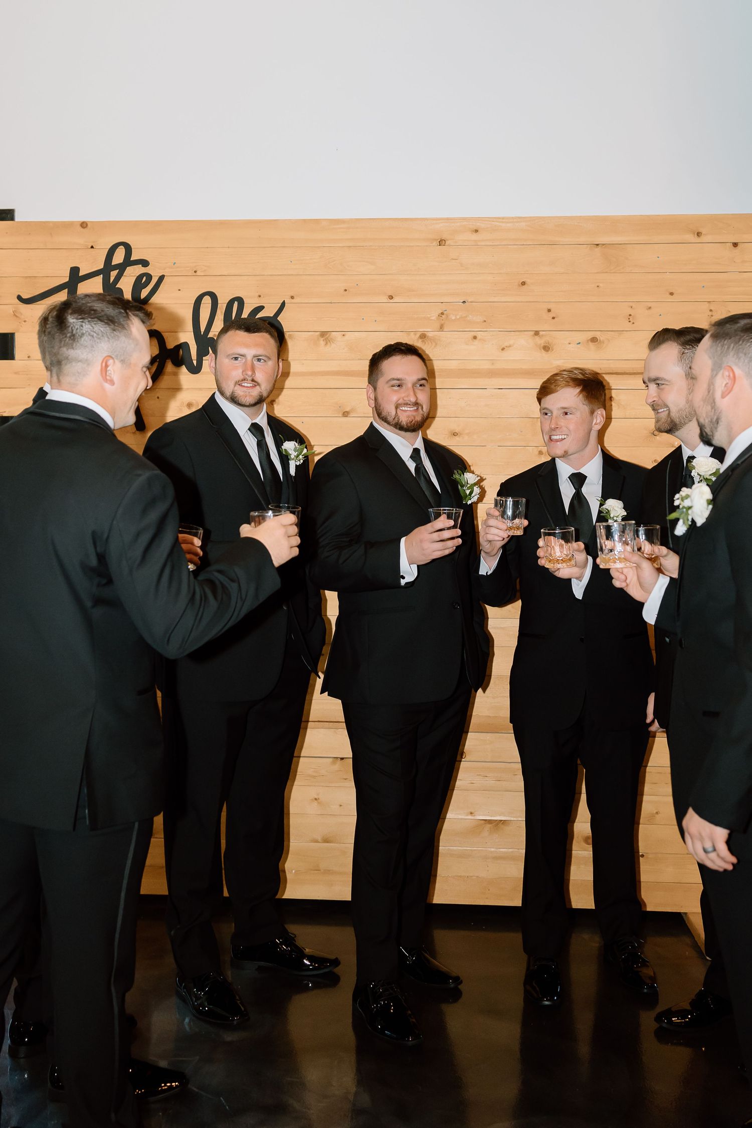 A group of groomsmen in black tuxedos share drinks and laughs during a wedding celebration.