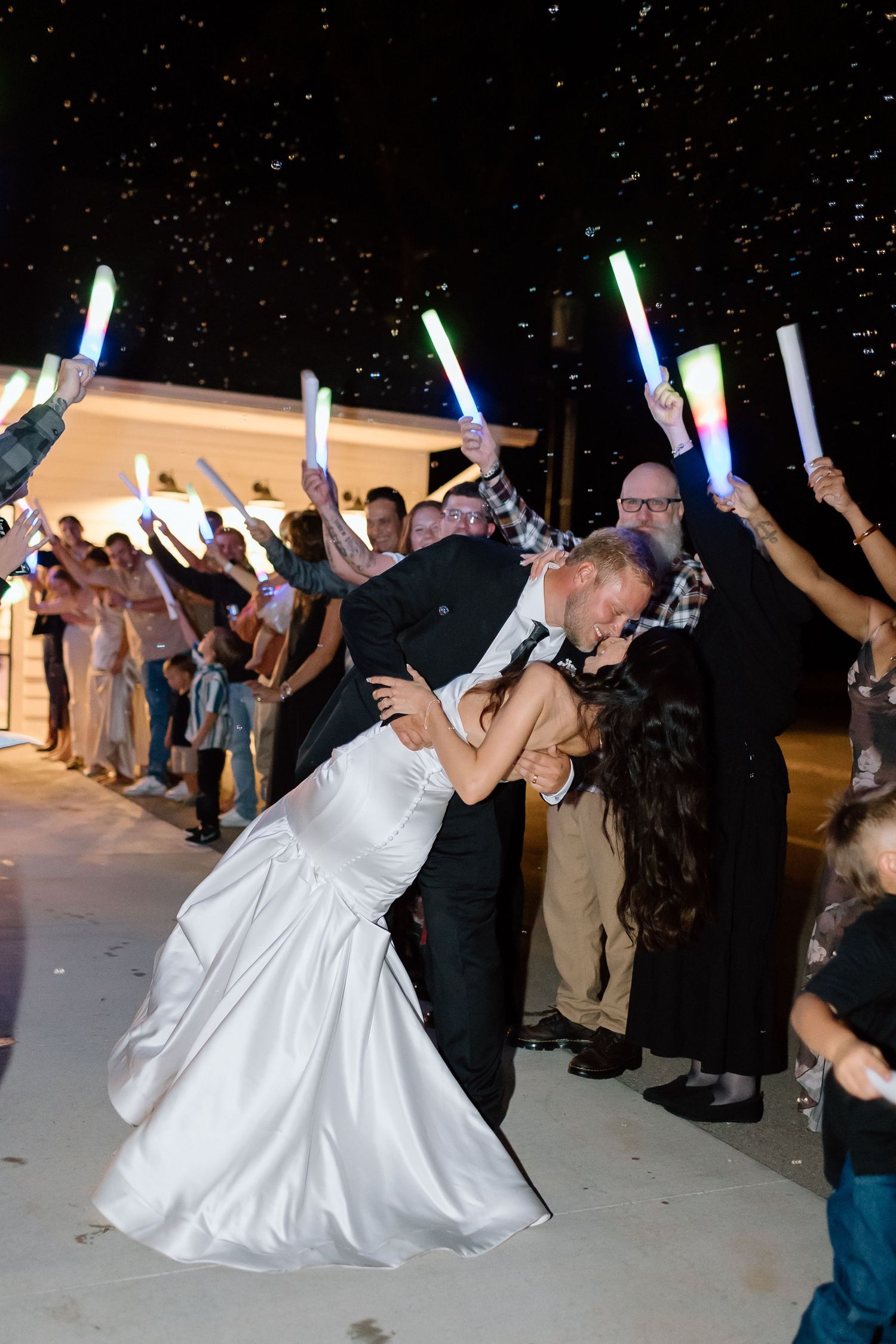 Wedding guests hold glowing light sticks during a nighttime sparkler exit for the newlyweds.