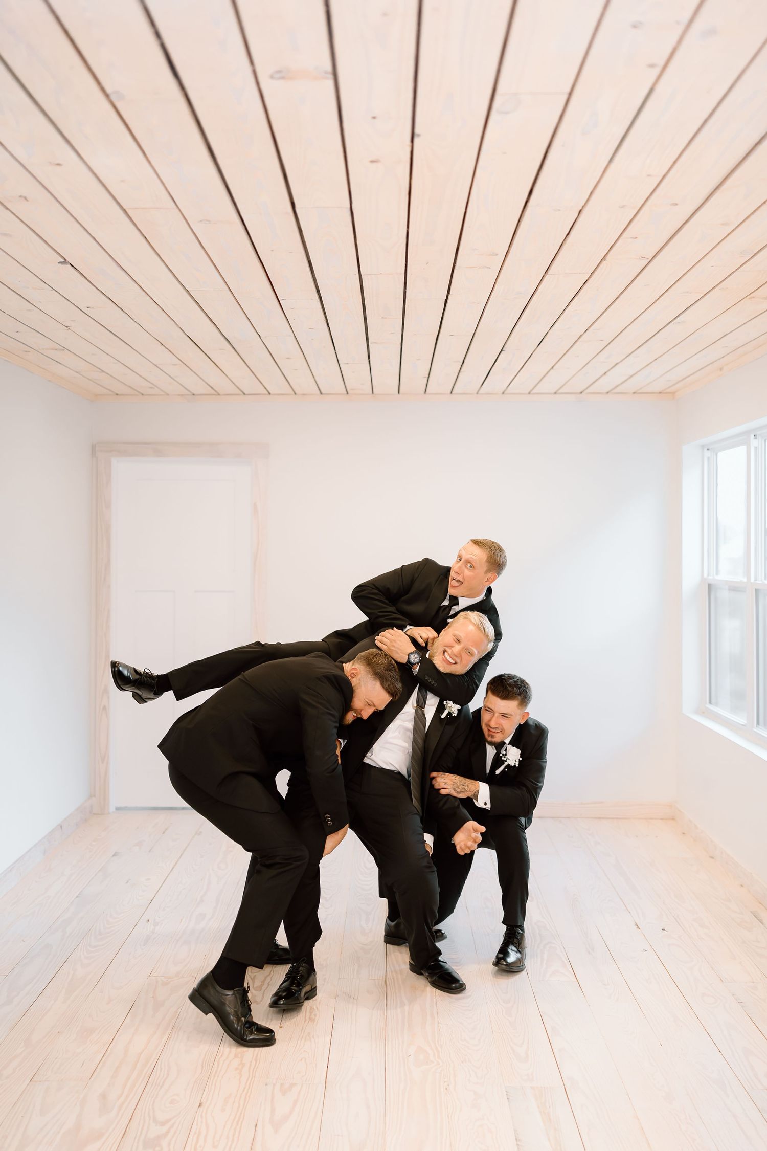 Three men in black suits playfully pose in sequence across a minimalist white room with wooden plank ceiling.