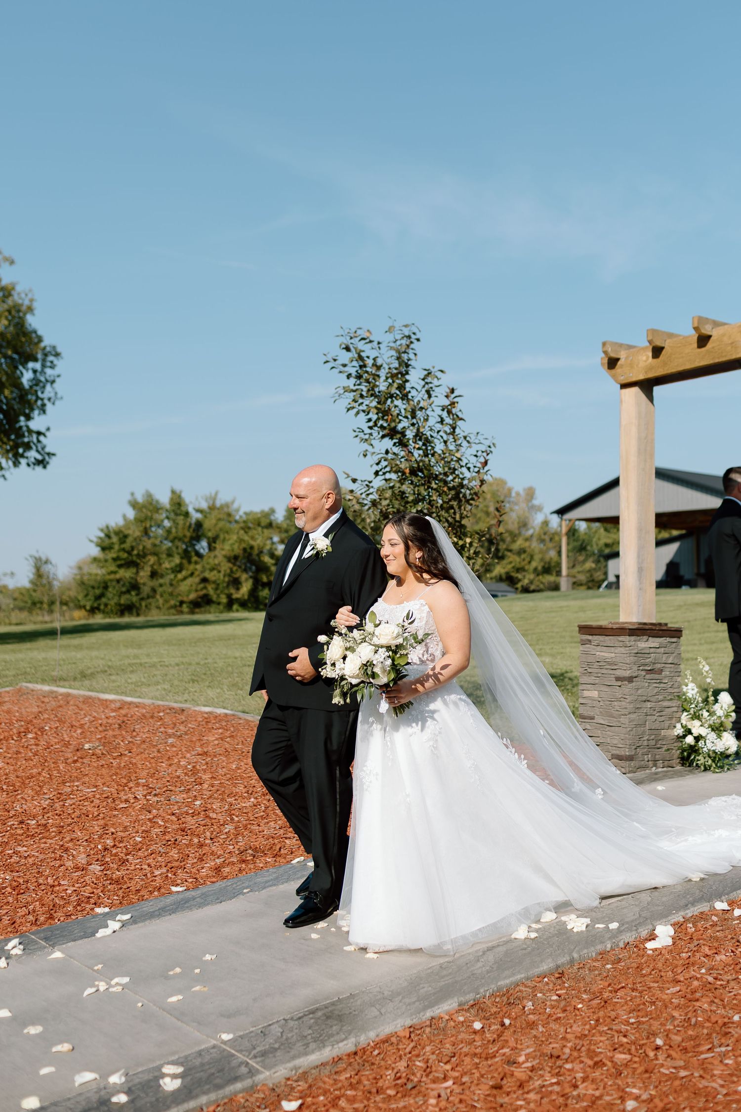 A bride in a white gown and veil walks down an outdoor path with an escort under a blue sky.