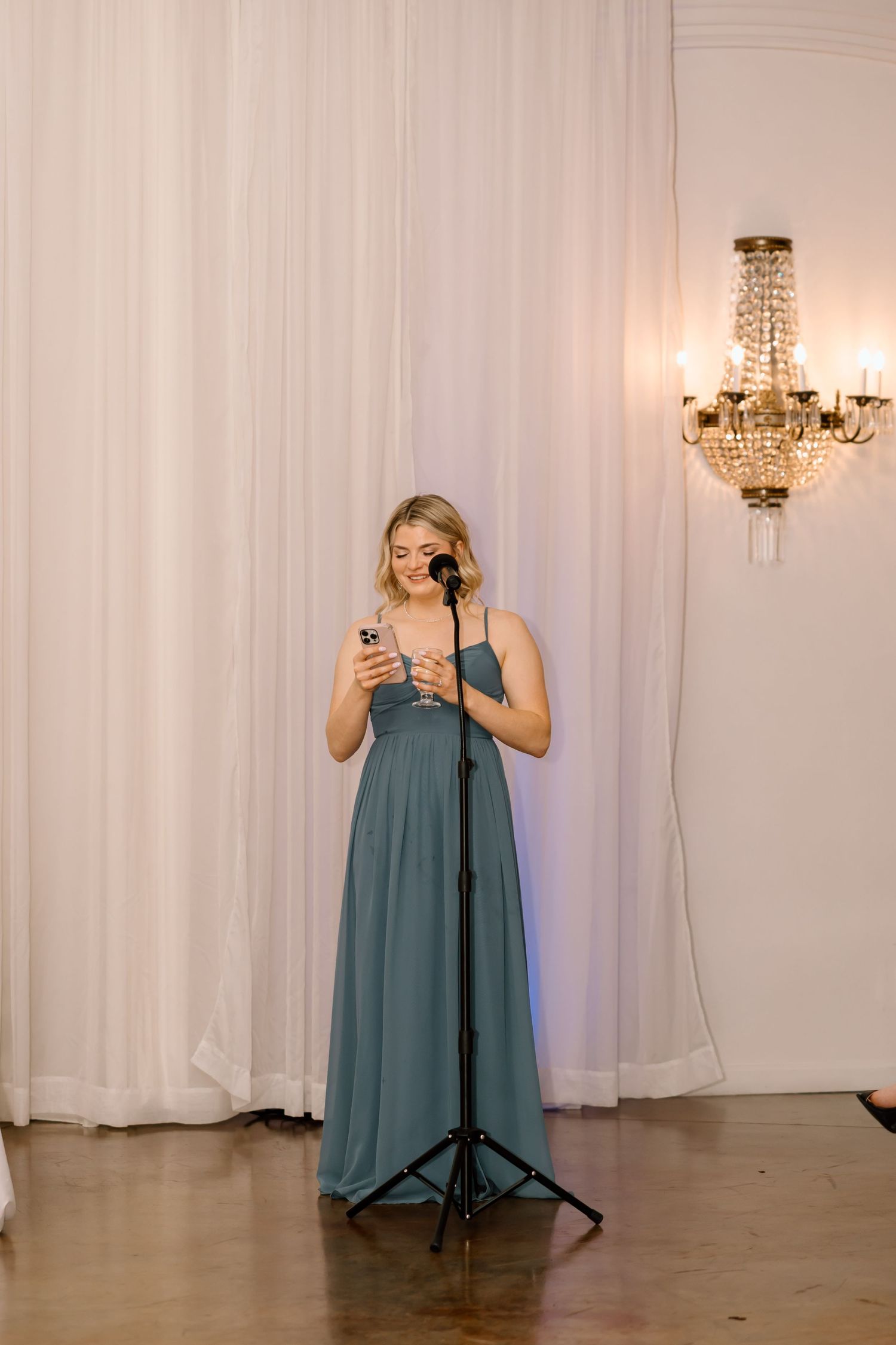 Someone in a dusty blue bridesmaid dress stands in an elegant venue with a crystal chandelier while checking their phone.
