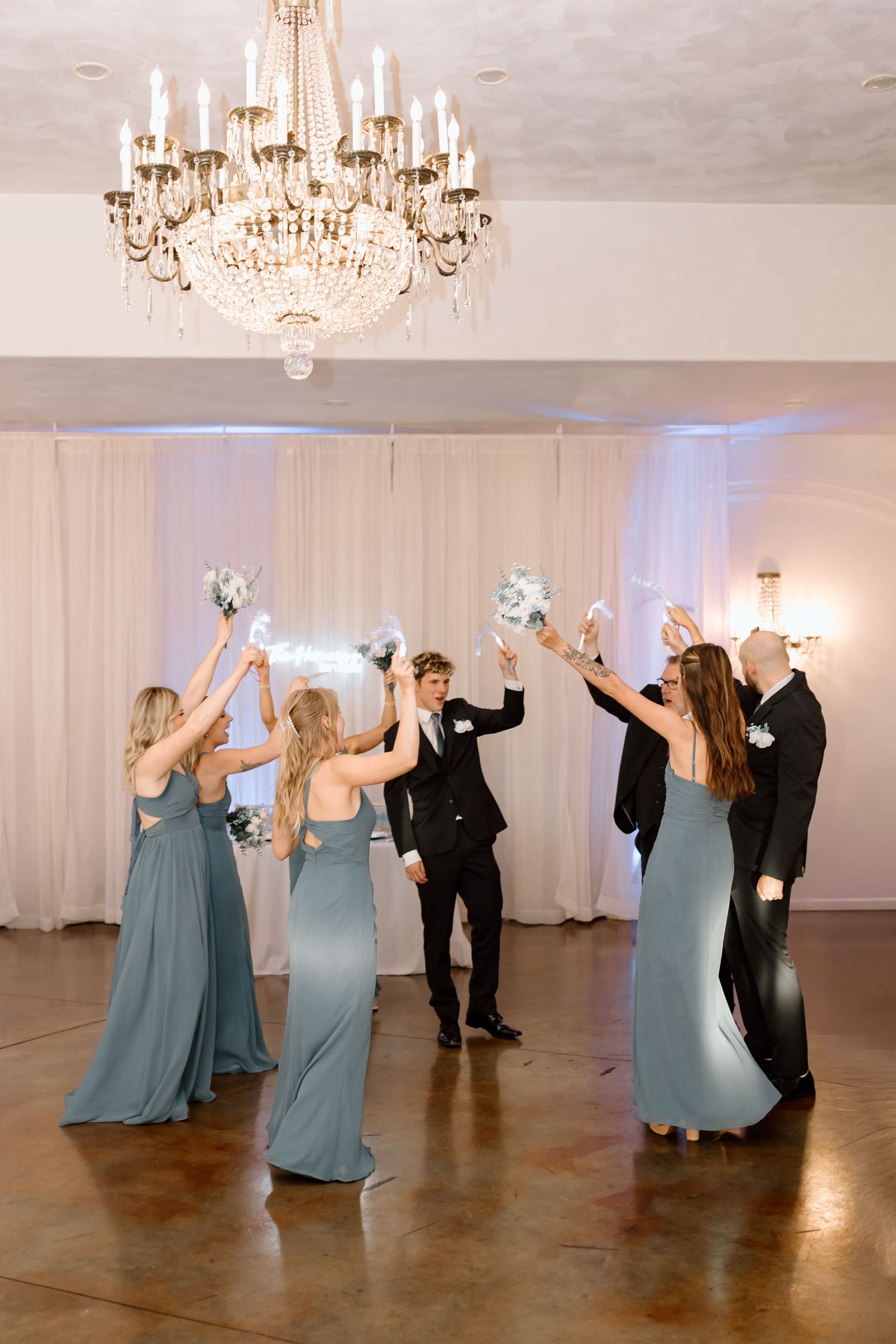 Wedding guests in dusty blue dresses dance in sequence across a ballroom floor with elegant chandeliers overhead.