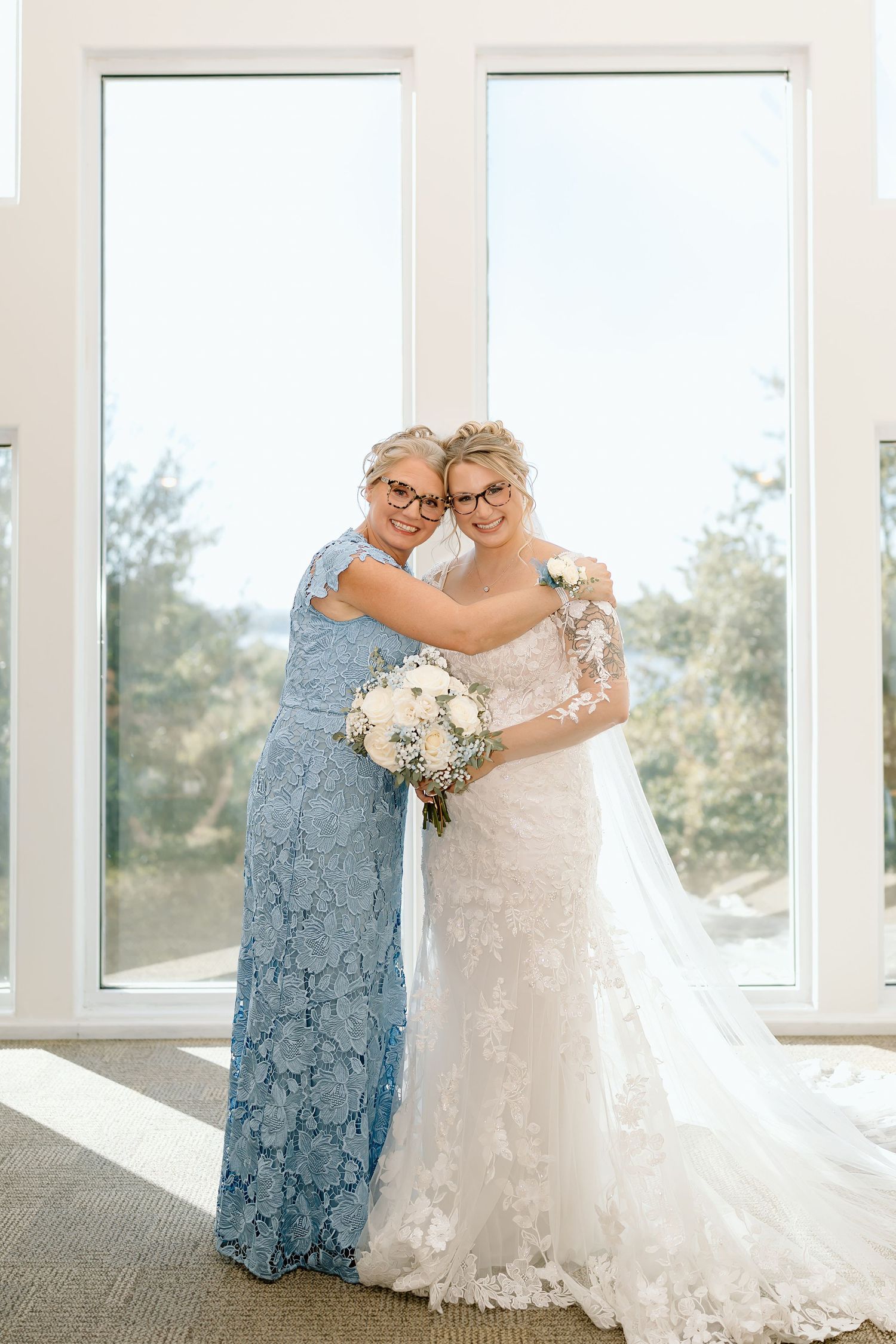 Two people embrace in a sunlit room, wearing a blue lace gown and white wedding dress while holding a white bouquet.