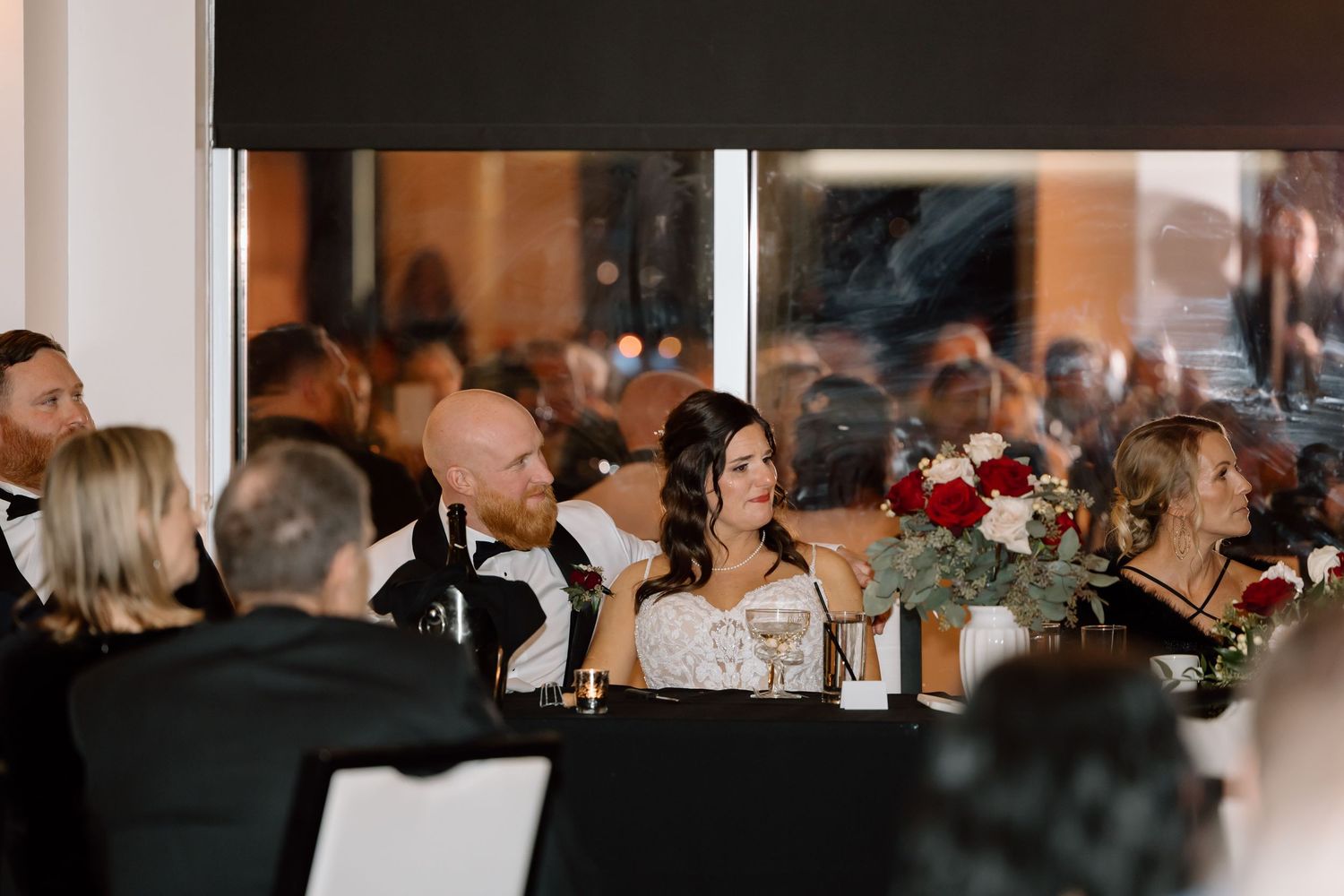 Wedding reception guests seated at formal dinner tables with floral centerpieces in an elegant indoor venue.