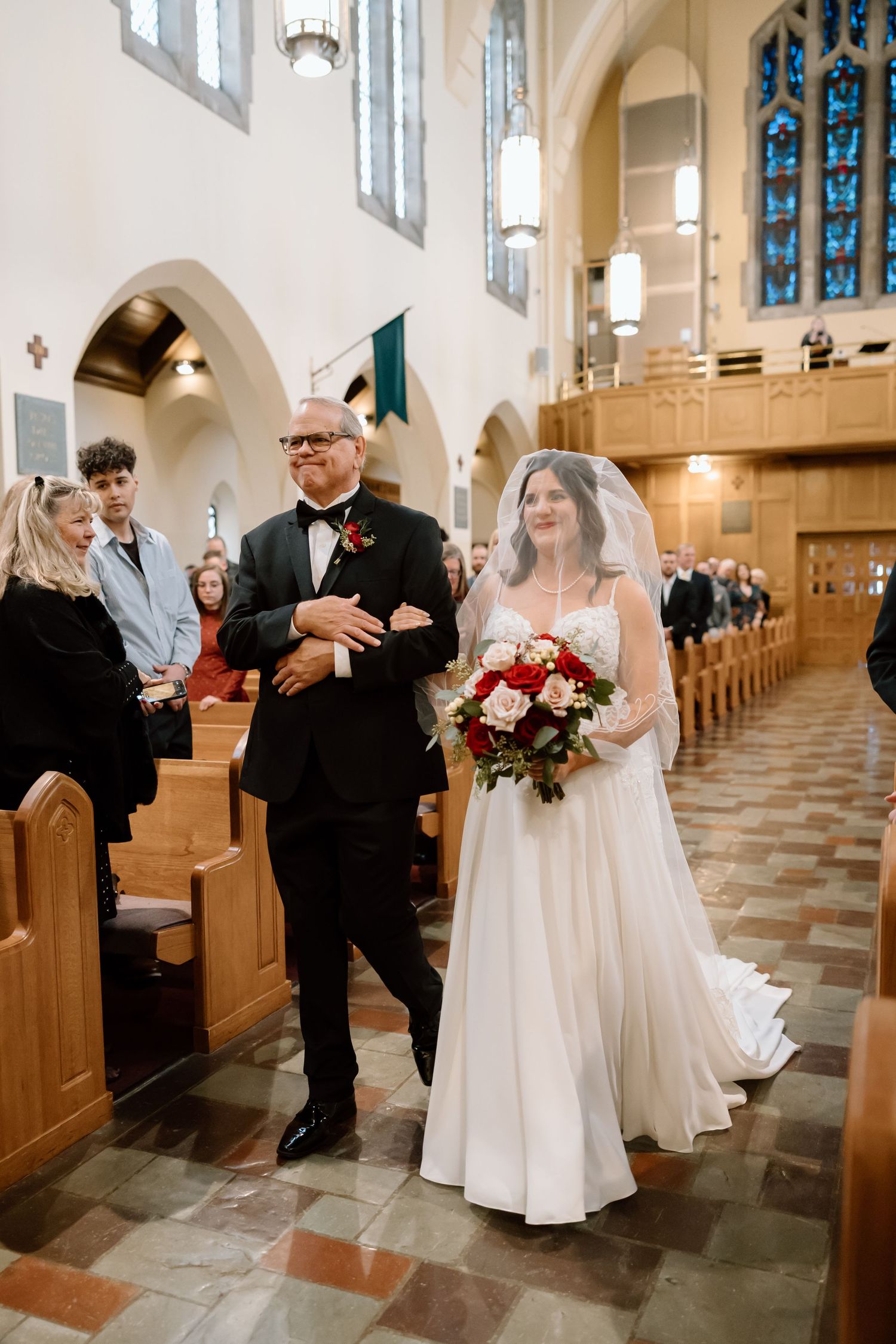 Traditional church wedding ceremony with bride in white gown holding red bouquet walking down aisle with escort.