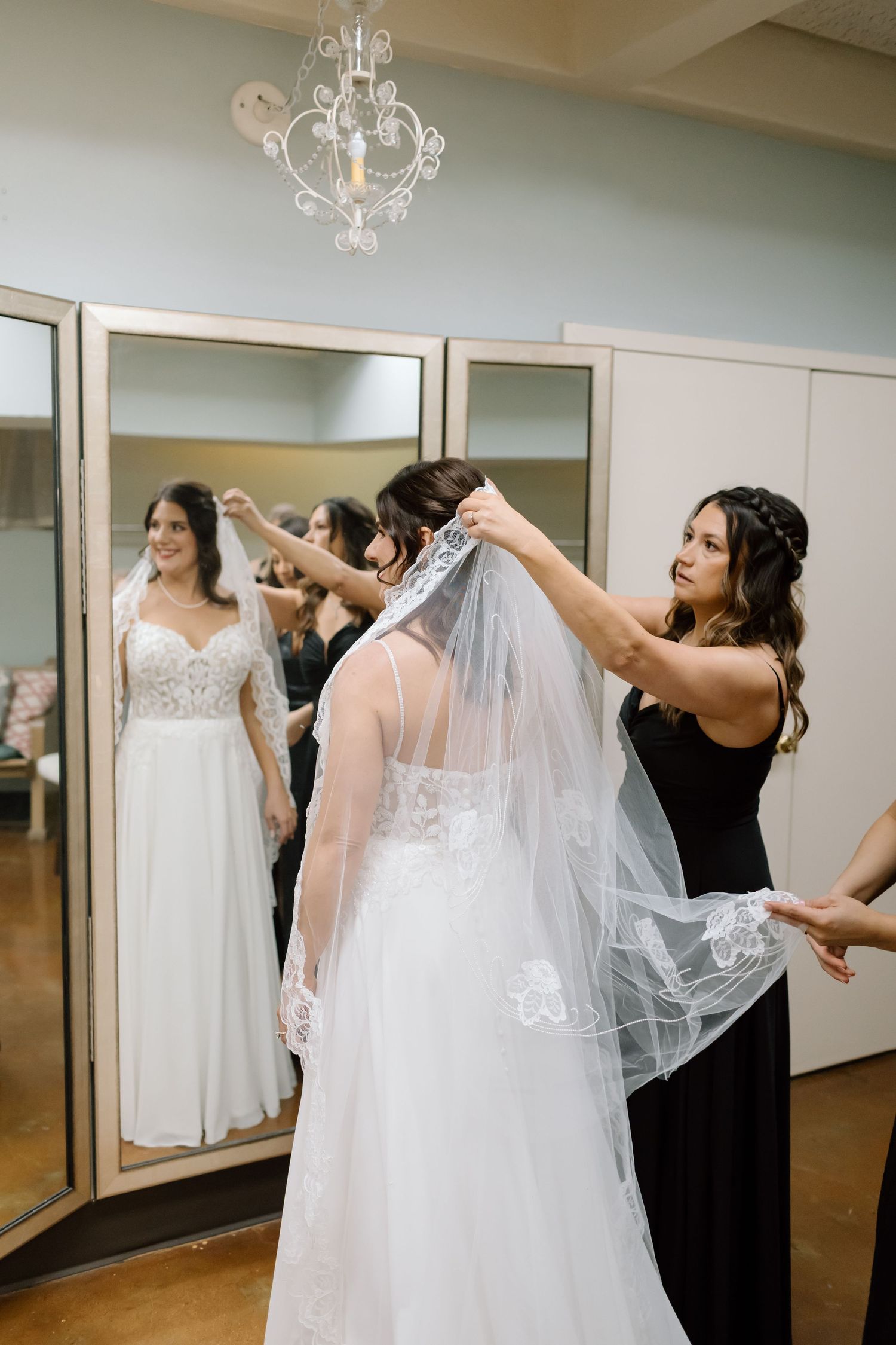 A bride getting help with her wedding veil in a bridal suite with mirrors and chandeliers.