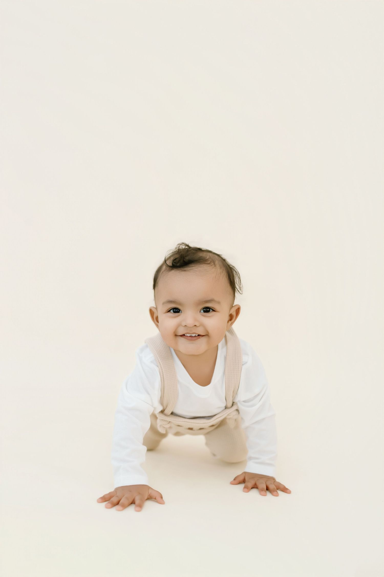 A baby in white clothing crawls and smiles on a light colored background.