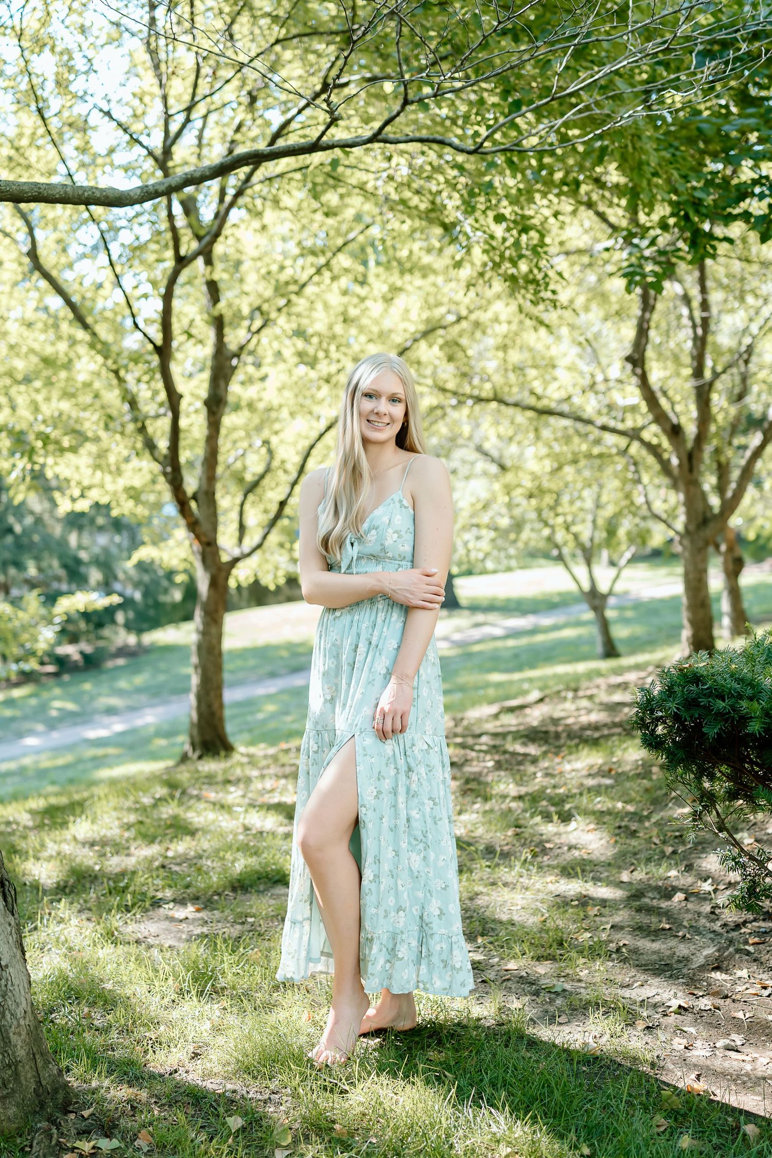 A light floral dress flows in the sunlight as someone poses gracefully among blooming trees in a peaceful garden setting.