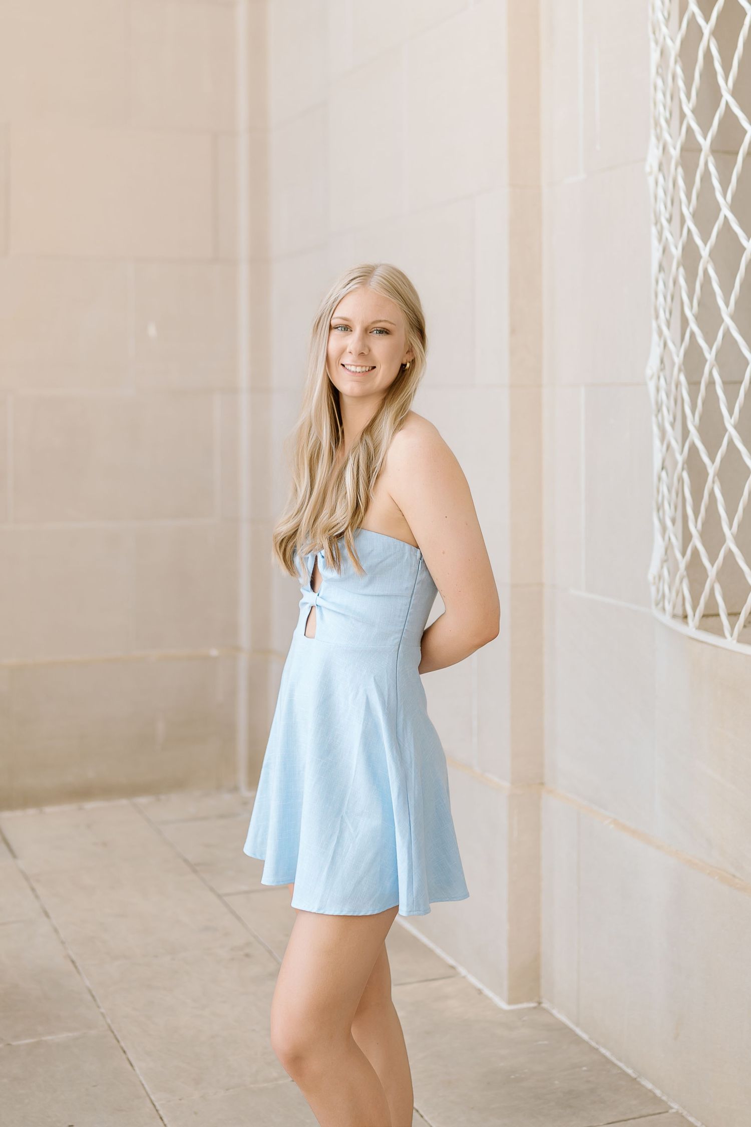 Someone poses in a light blue summer dress against a beige wall with decorative lattice details.