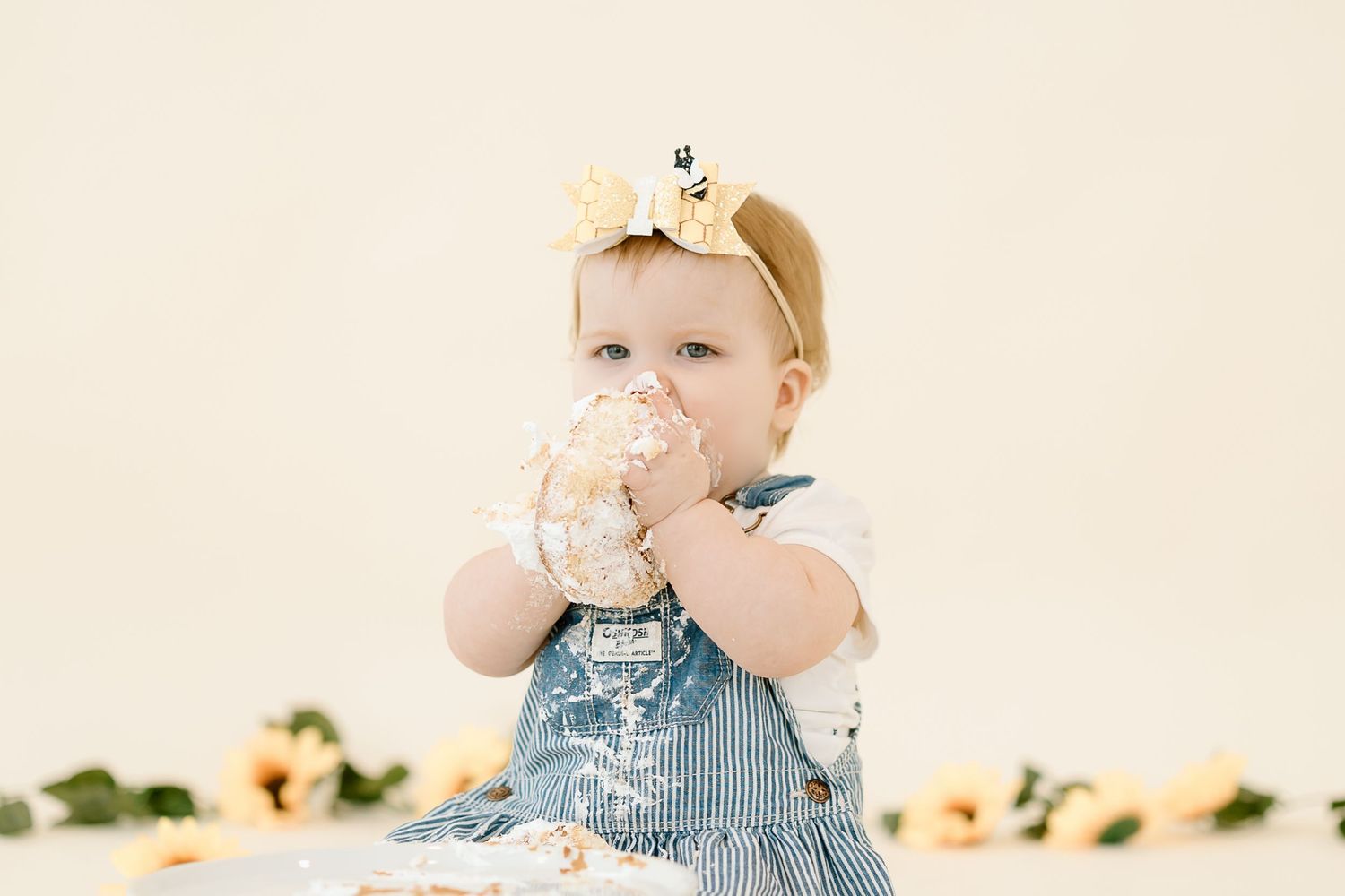 Baby in blue striped overalls drinks from a glass cup while sitting at a wooden table with colorful fruit.