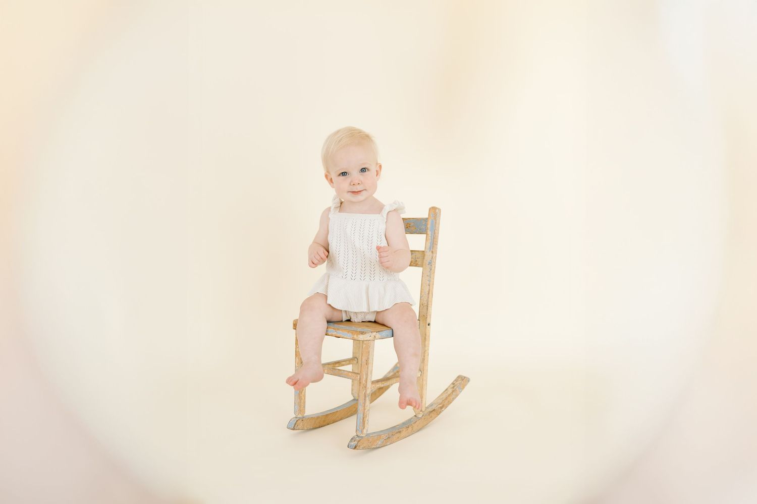 A baby in white romper stands next to vintage rocking chair with white balloon in studio setting.