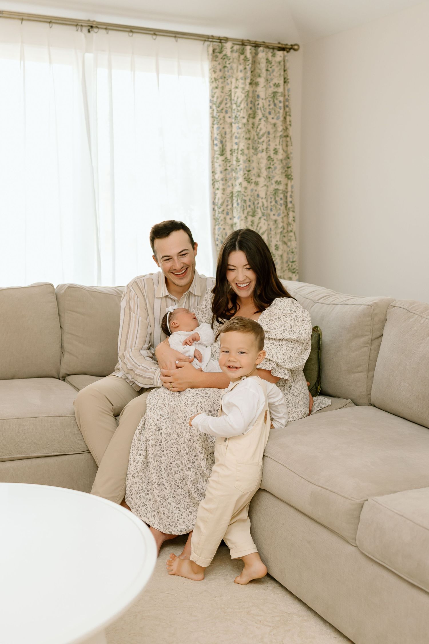 A family sits together on a beige sectional sofa in a bright, minimalist living room with floral curtains.