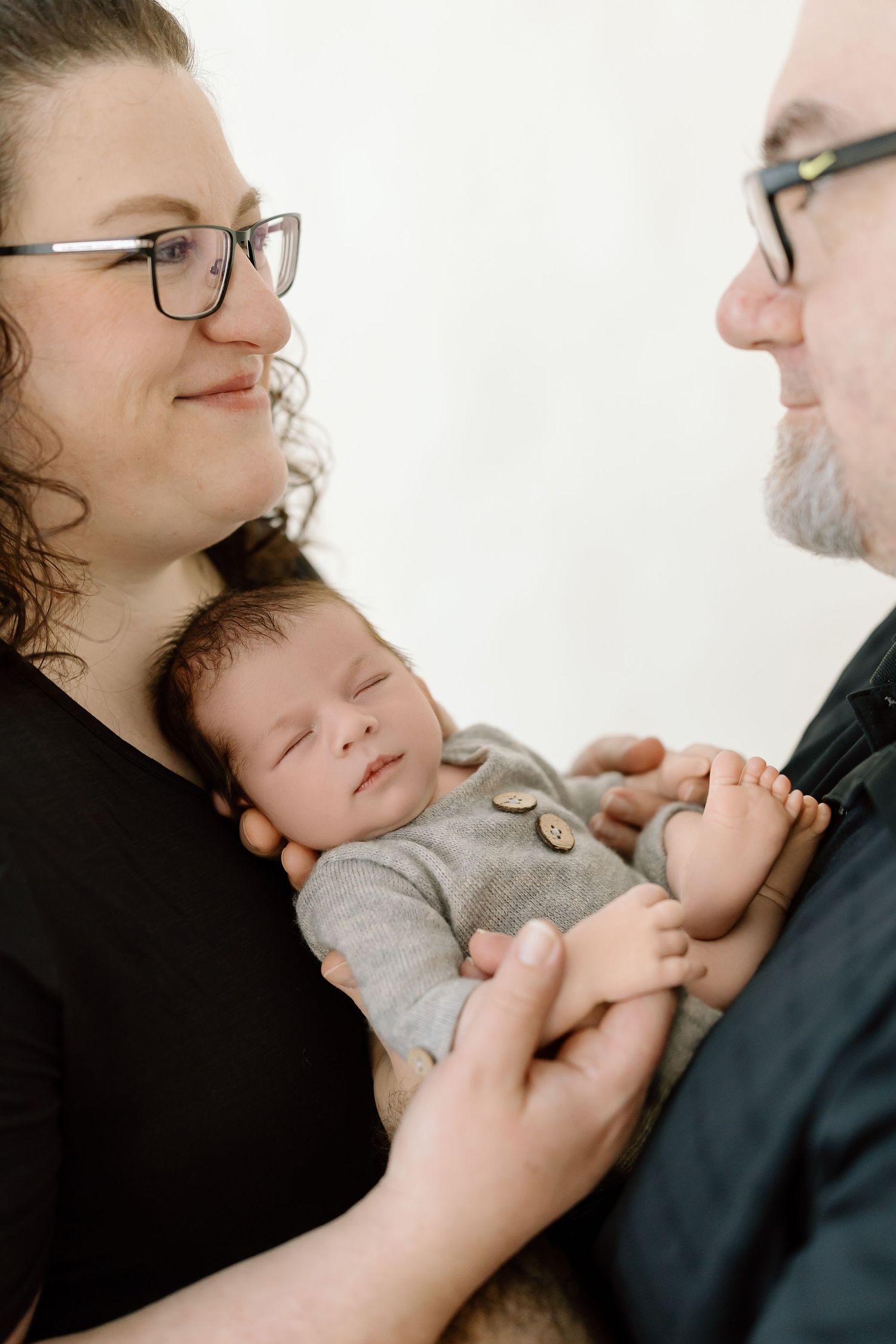 A sleeping baby in a gray onesie is cradled between two adults wearing glasses in an intimate family portrait.