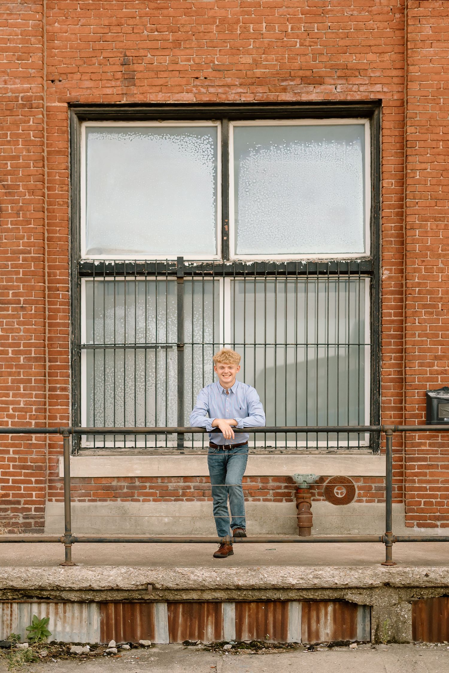 A person sits alone on a bench outside a brick building with large windows and metal railings.