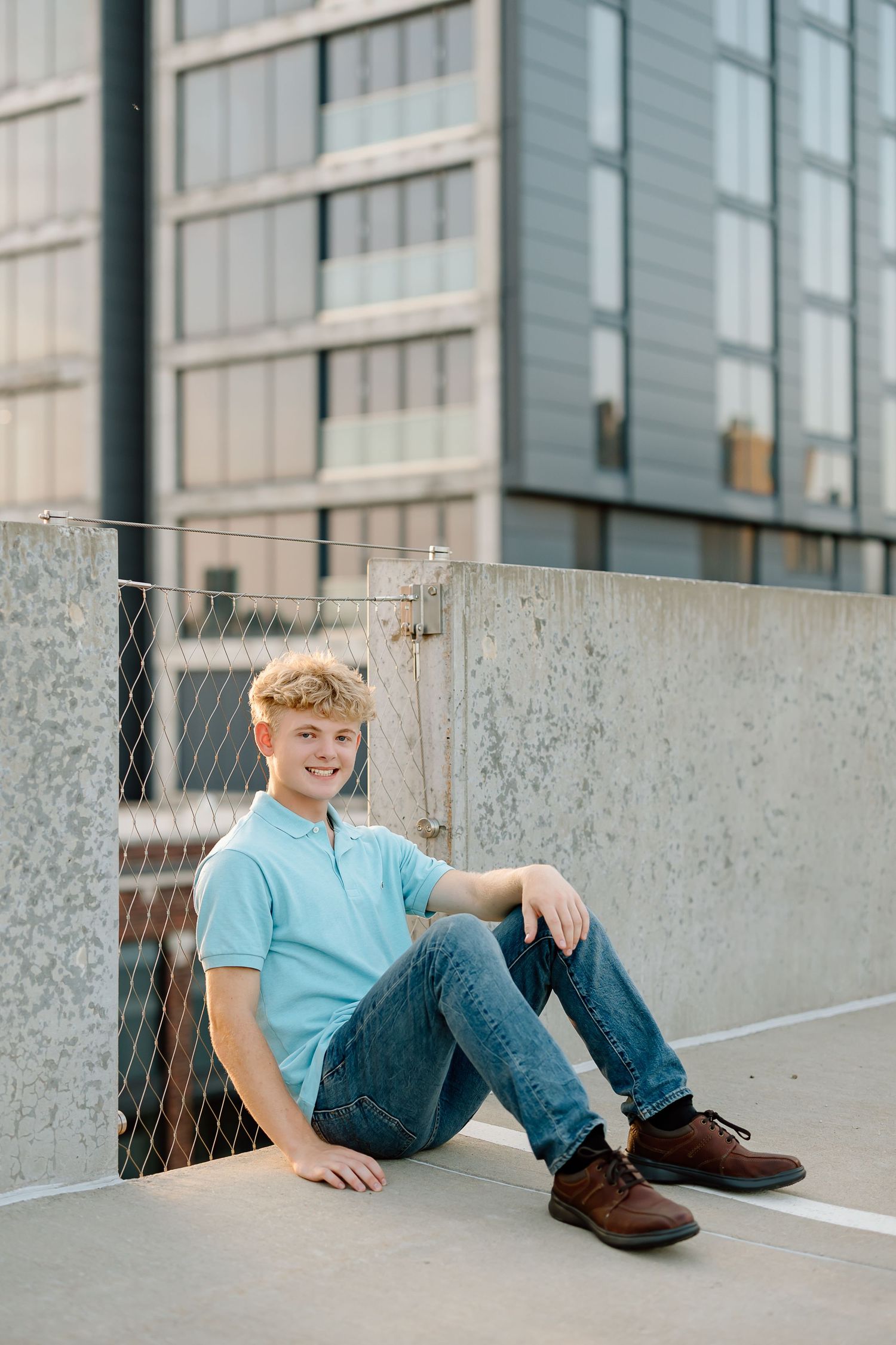 A person in casual attire sits on a concrete surface with modern glass buildings visible in the urban setting.
