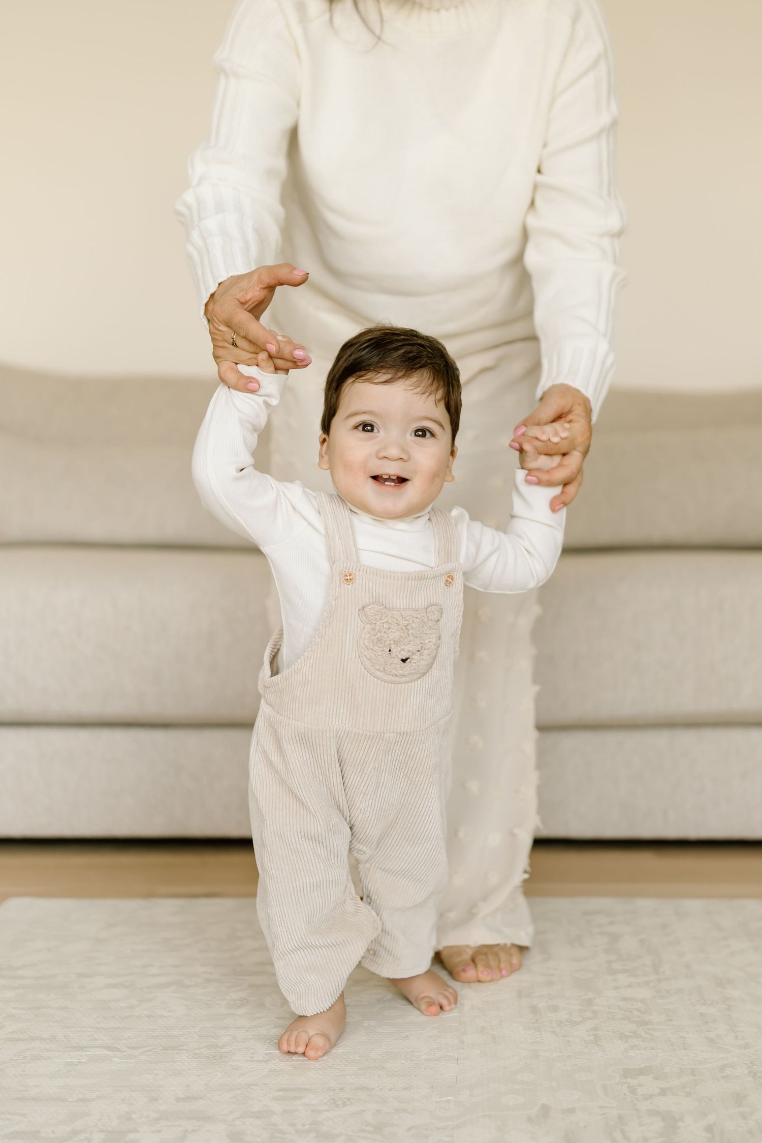 A baby in white overalls takes first steps while holding hands with an adult in a bright indoor setting.