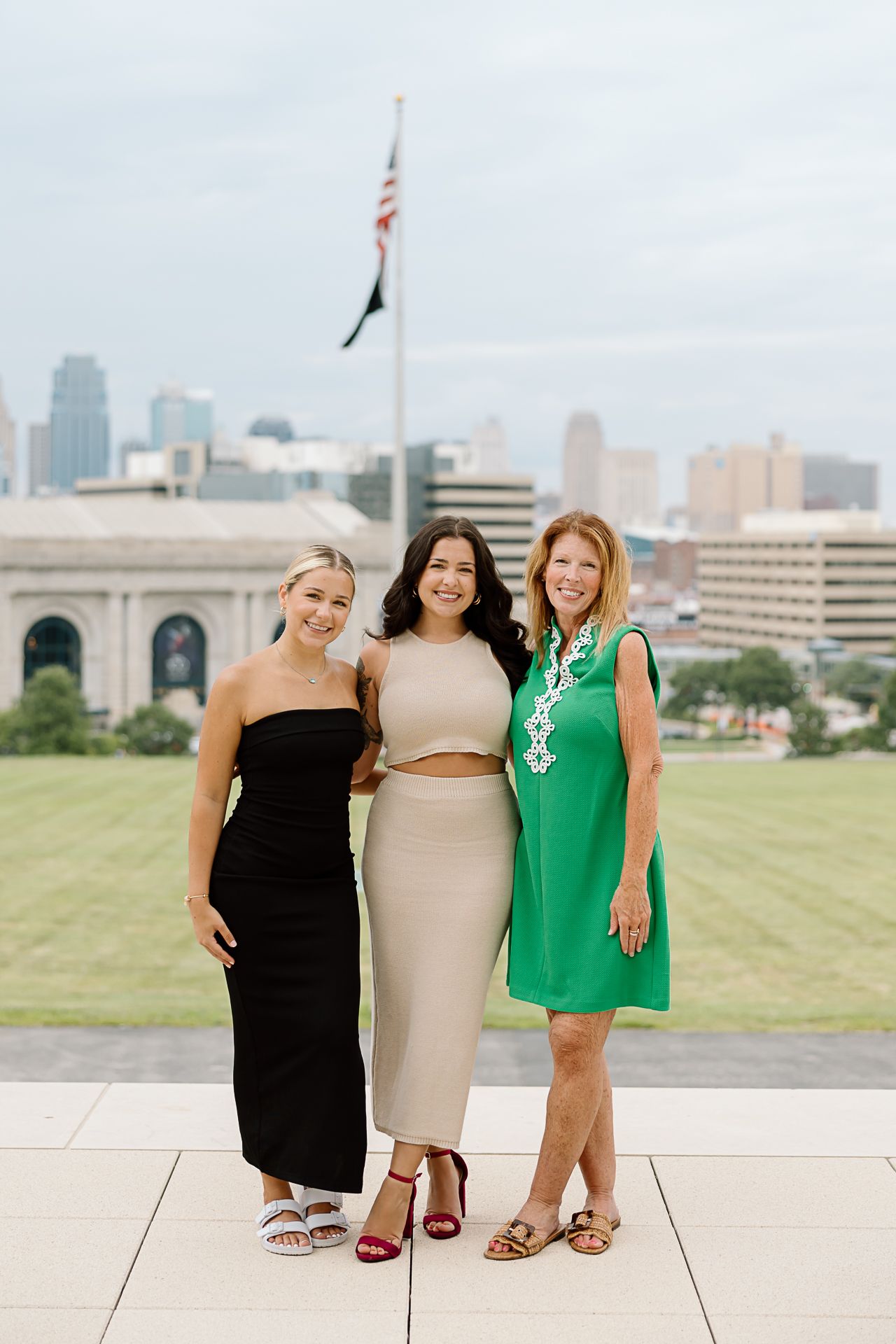 Three friends pose together outdoors wearing coordinated black, beige and green dresses with a city skyline in background.