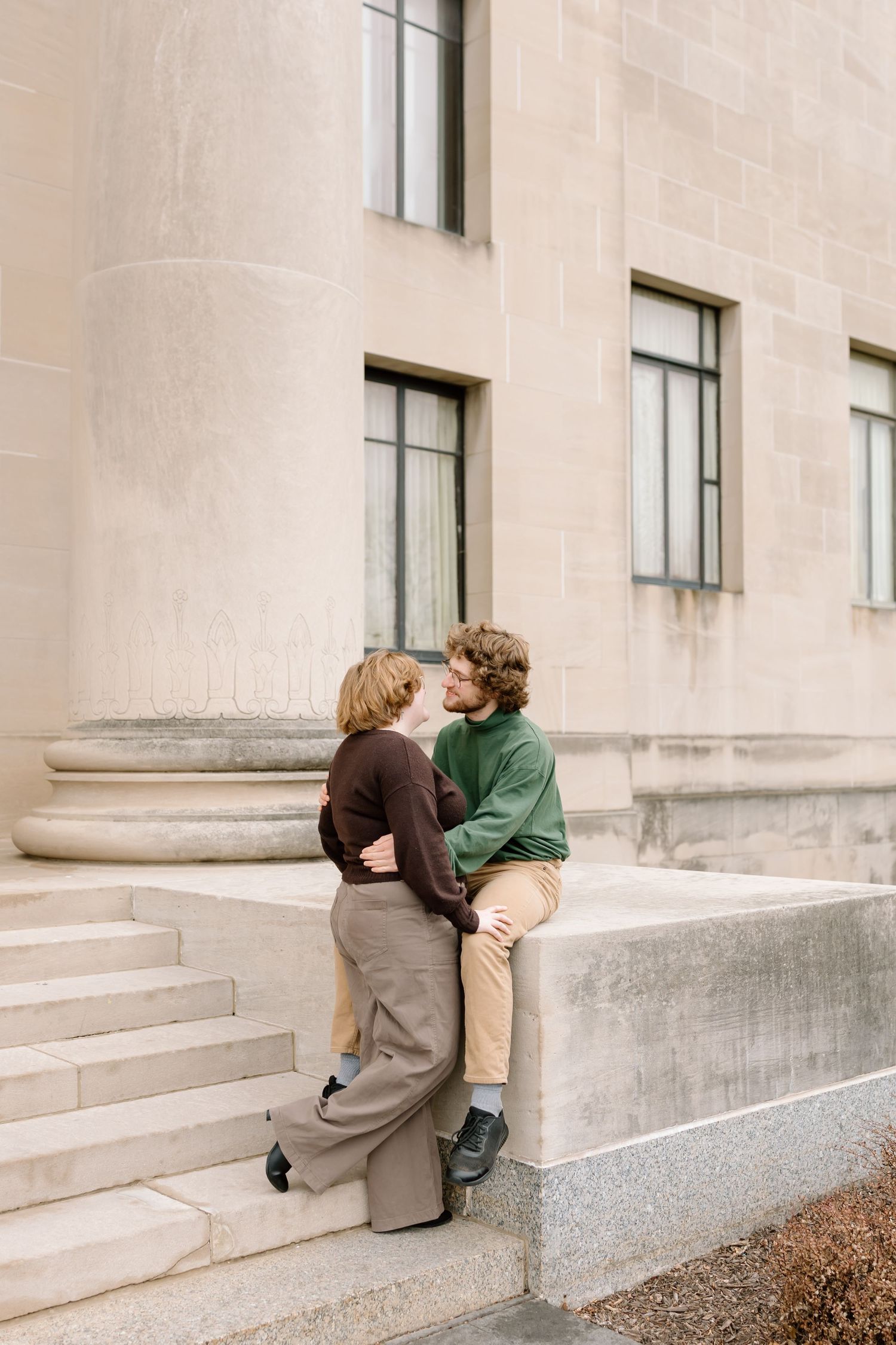 Two people share an intimate moment sitting on concrete steps against a light-colored building wall.