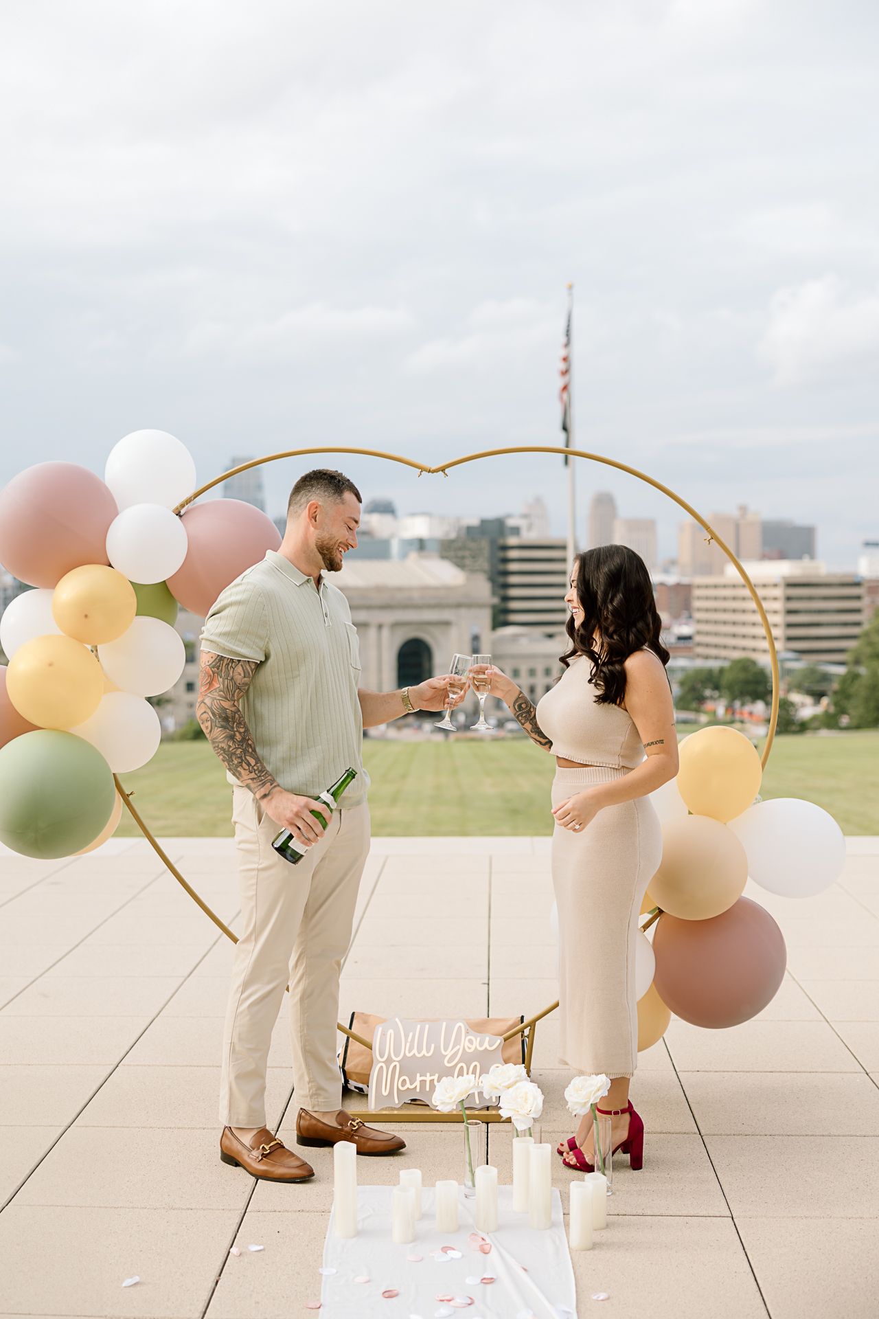A romantic rooftop proposal scene decorated with pastel balloons and signage against a city skyline backdrop.