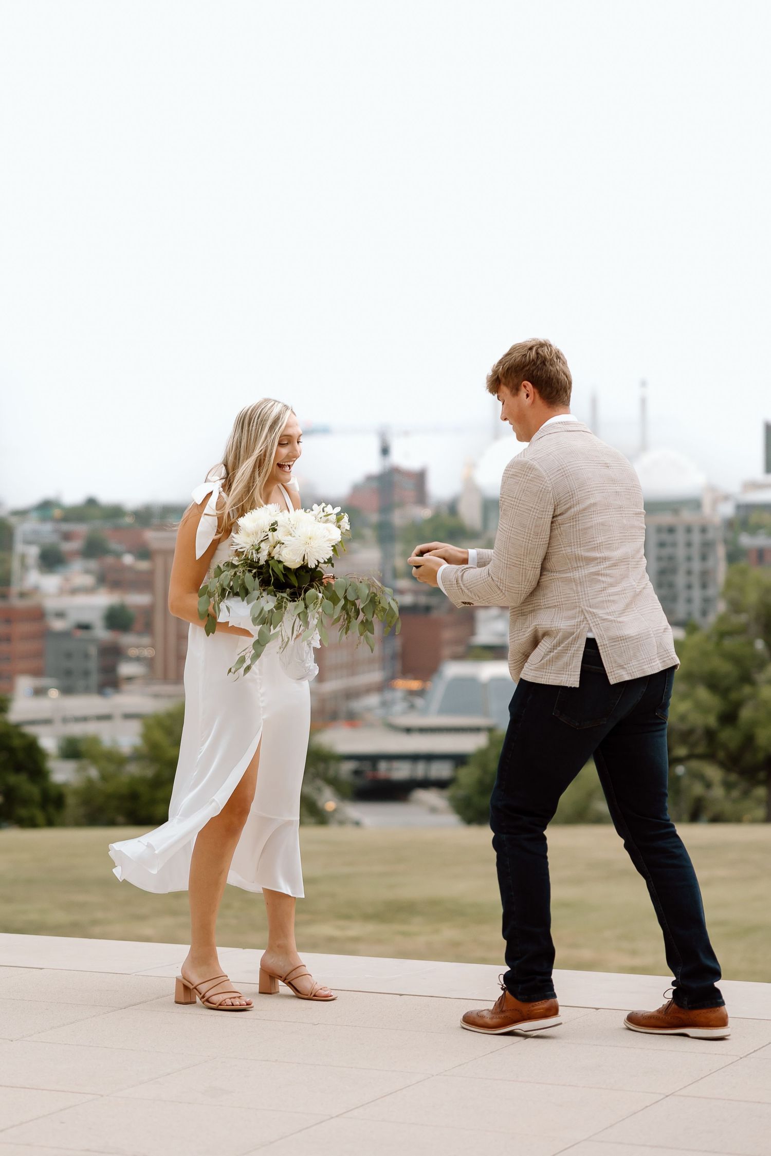 A couple in white and beige attire playfully pose for photos on a hilltop overlooking a cityscape.
