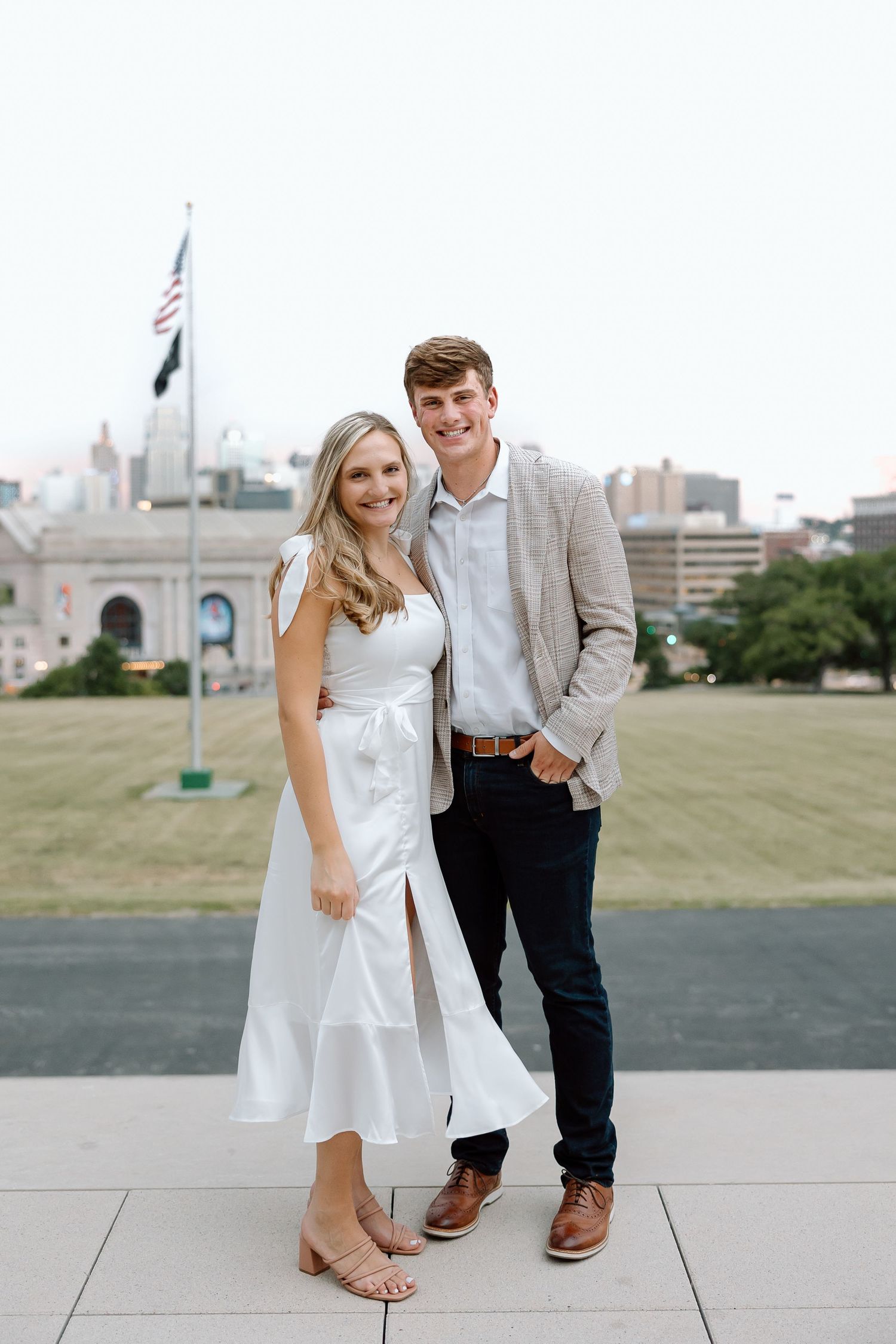 A couple in formal attire poses together on a rooftop terrace with a city skyline in the background.