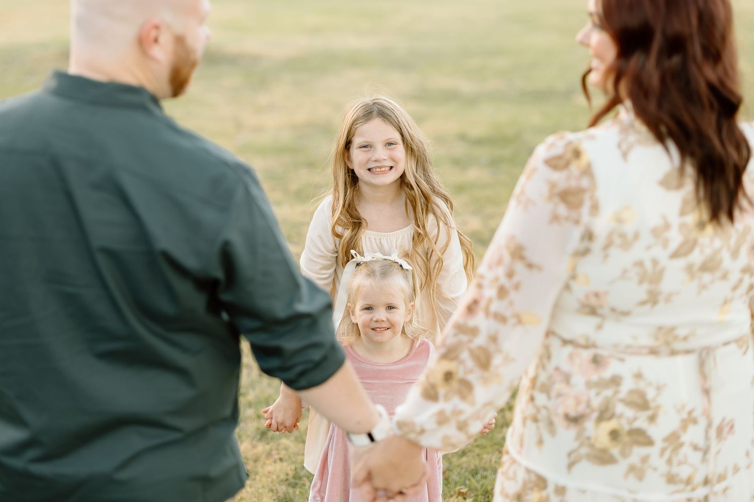 Family holding hands and smiling together in a sunny outdoor field during golden hour.