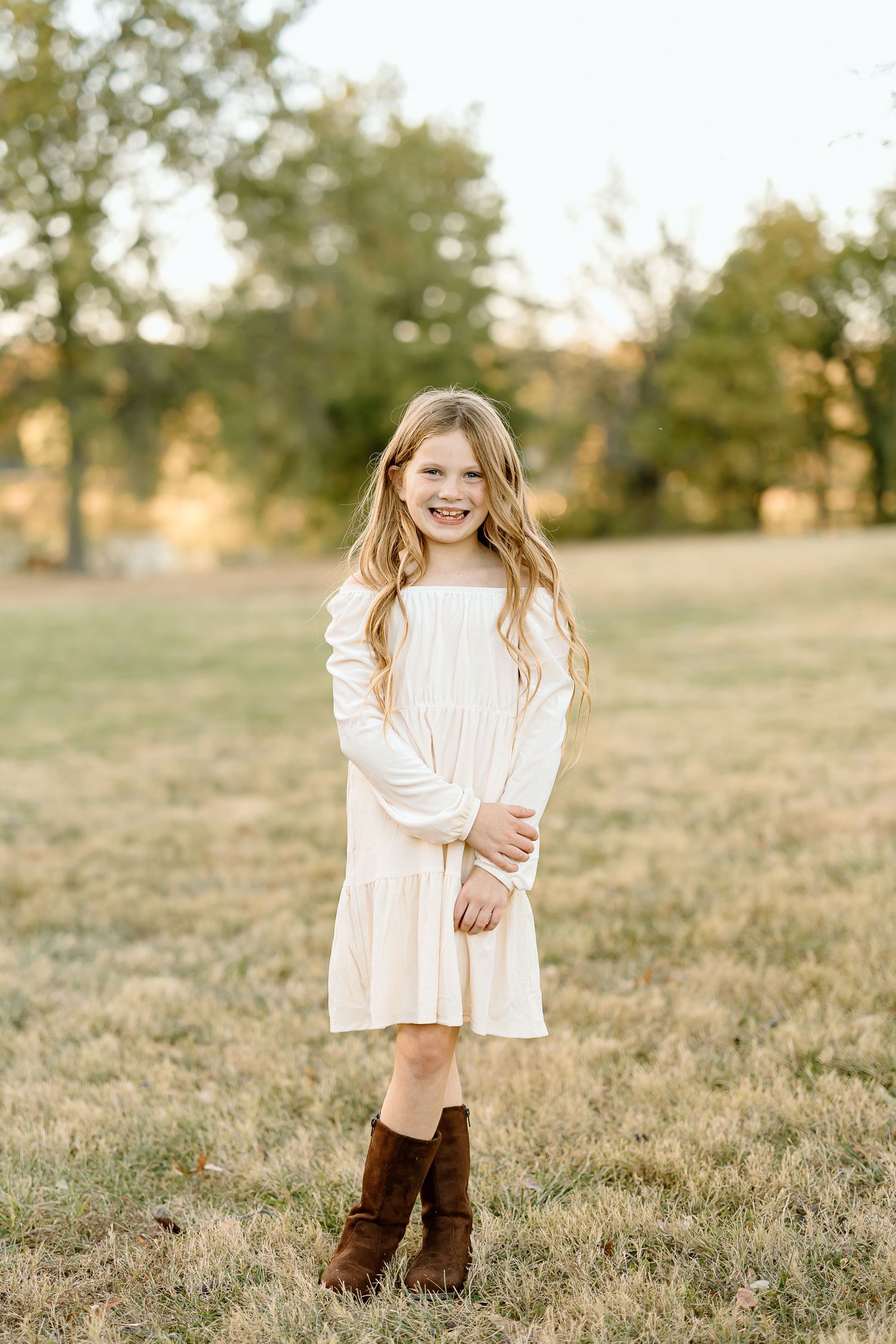 A child wearing a white dress and brown boots stands in a sunlit grassy field during golden hour.