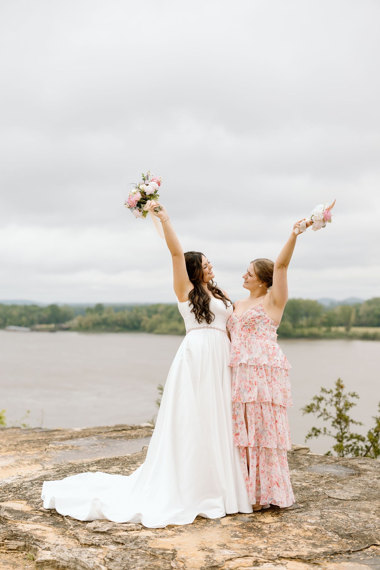 Two people in white and pink dresses sit together on a rocky cliff overlooking a scenic lake on an overcast day.