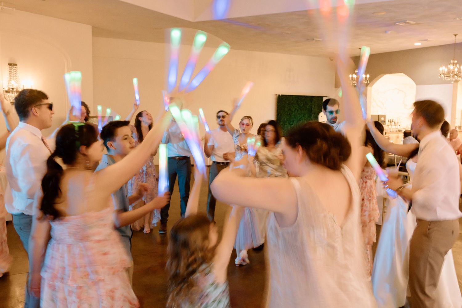 Wedding guests dance and celebrate on a crowded reception dance floor under colorful party lights.