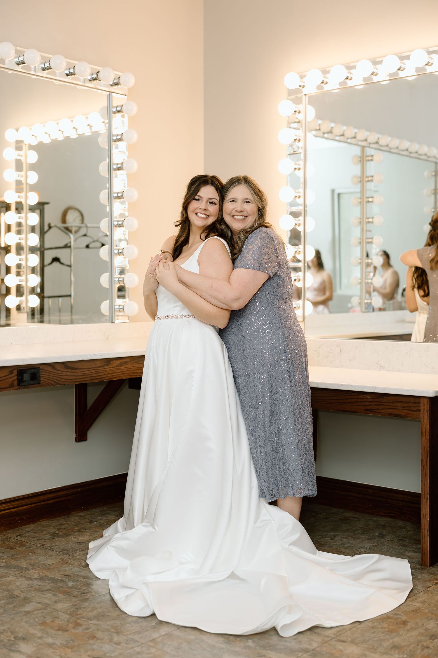 Two people pose together in a dressing room with illuminated vanity mirrors and wooden benches.