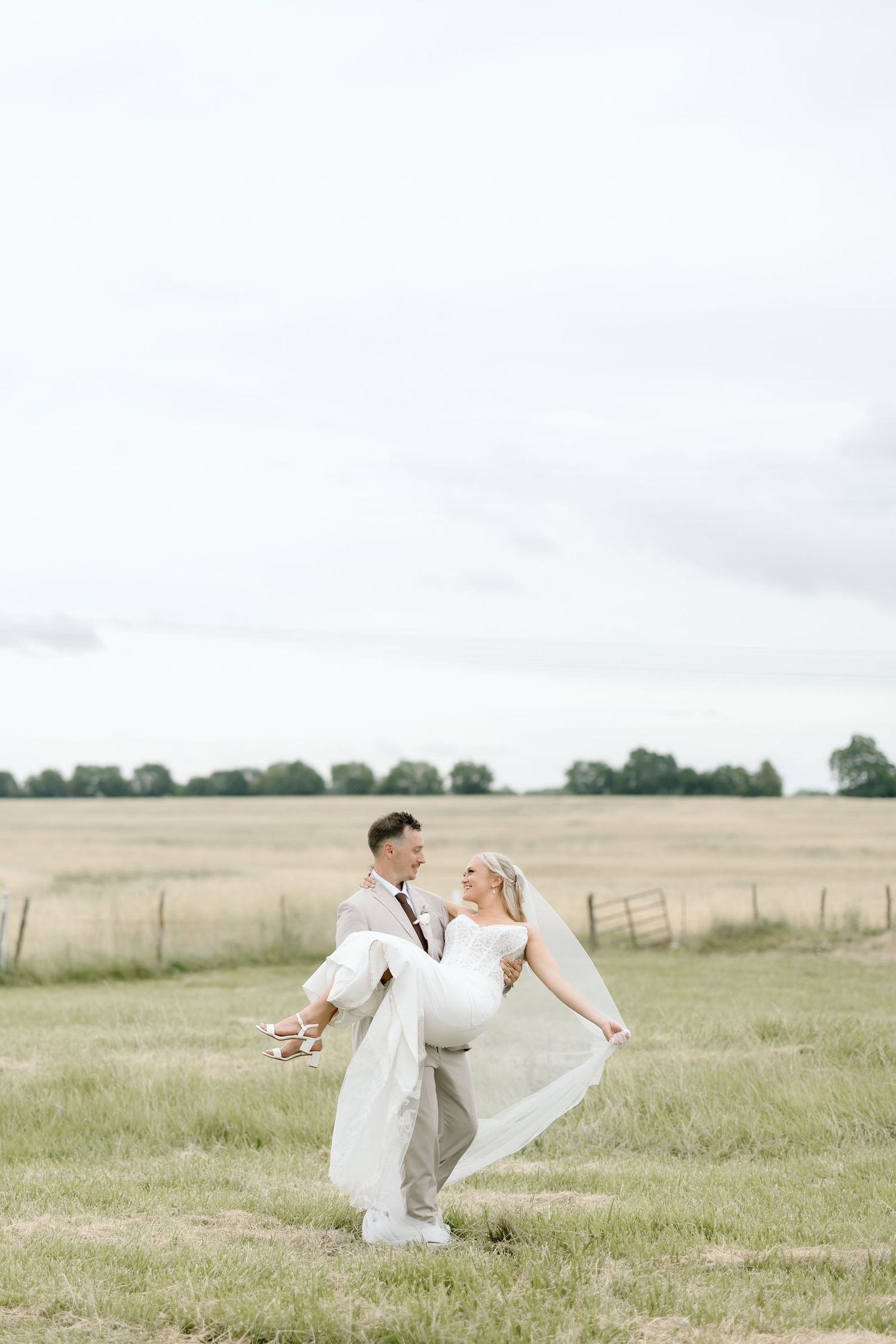 Groom lifting bride in white dress and veil in a golden field on their wedding day.