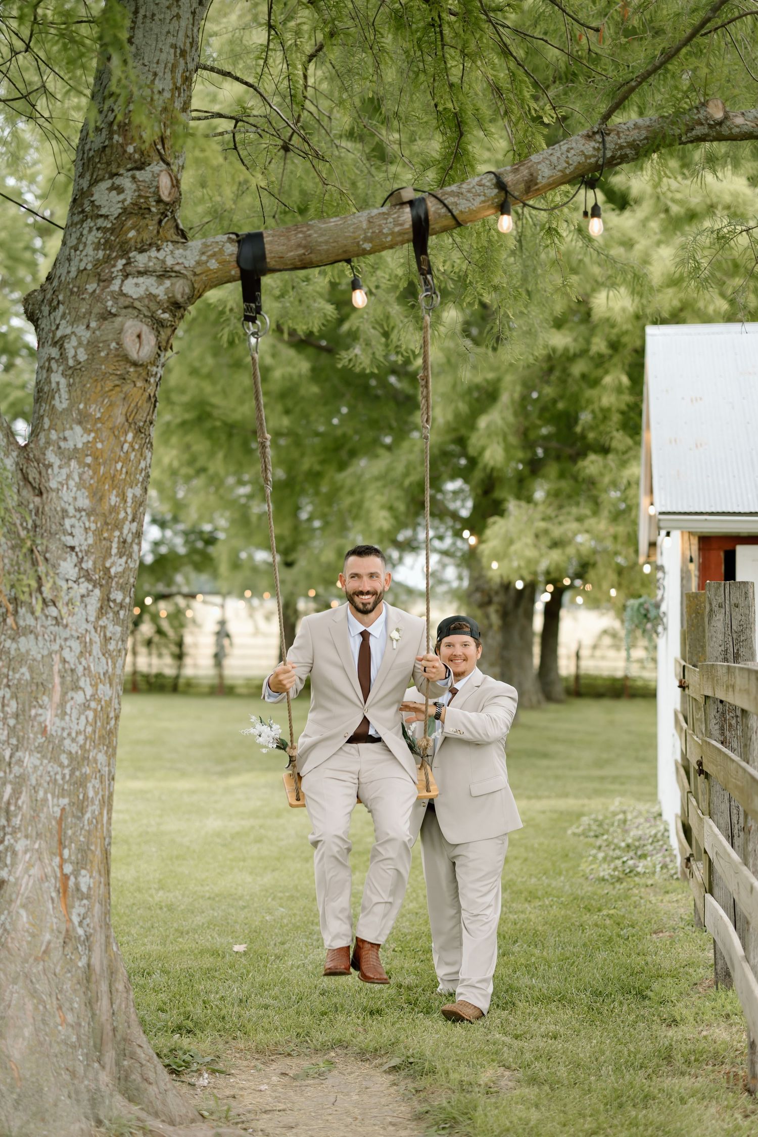 Two grooms in matching beige suits stand smiling by a rustic tree swing at an outdoor wedding venue.