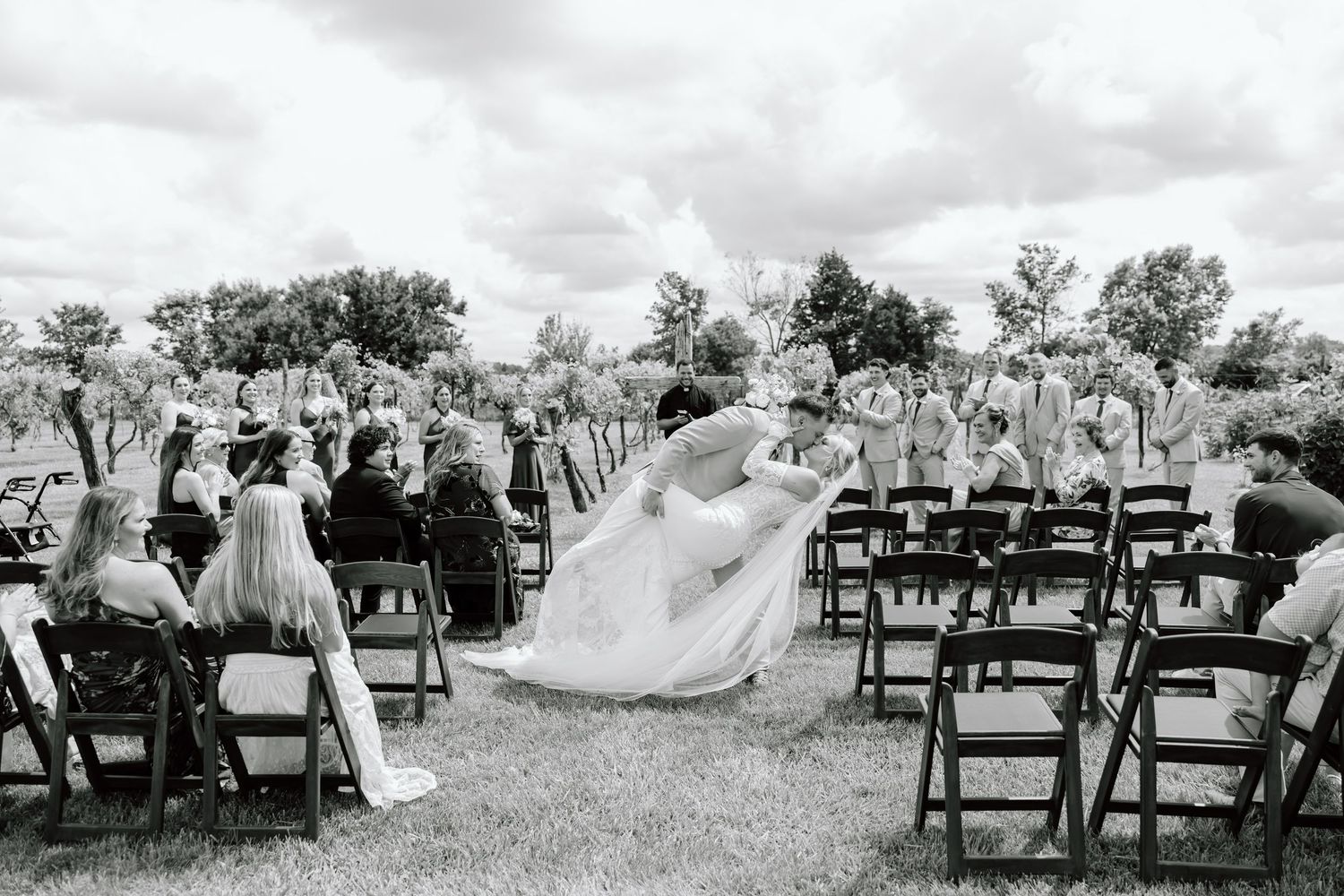 Black and white outdoor wedding ceremony with bride and groom at altar, guests seated in chairs on lush green lawn.
