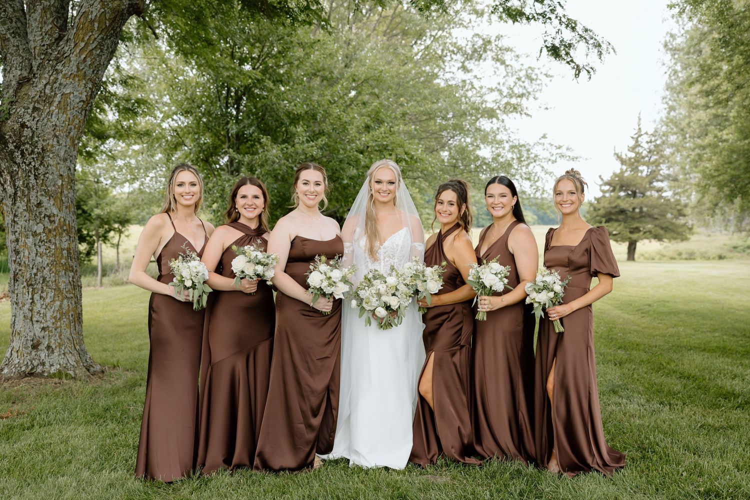 Bride in white gown poses with six bridesmaids in brown satin dresses holding white floral bouquets outdoors.