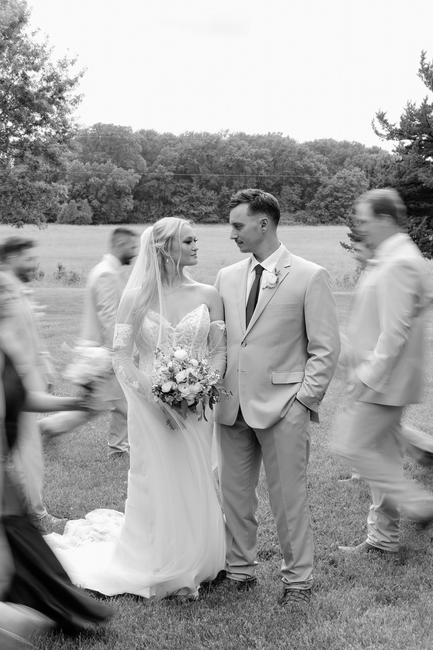 Black and white wedding photo of bride and groom standing still while wedding party moves around them outdoors.