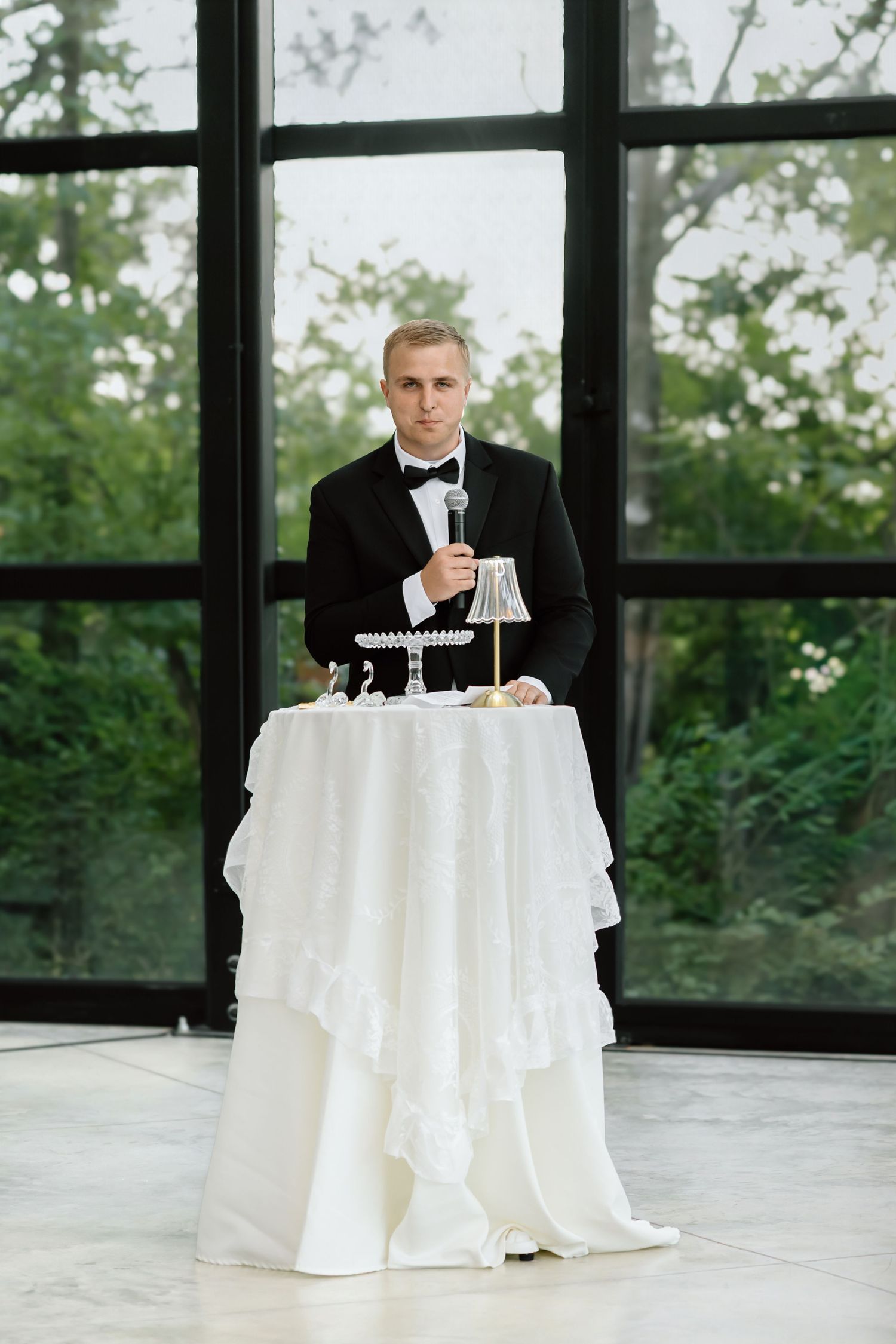 Elegant groom in black tuxedo and bow tie standing at white draped table during wedding reception indoors.