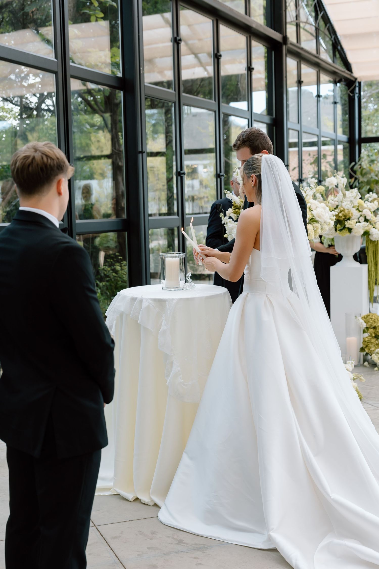 Bride in white gown lights unity candle at wedding ceremony with groom watching near floral arrangements.