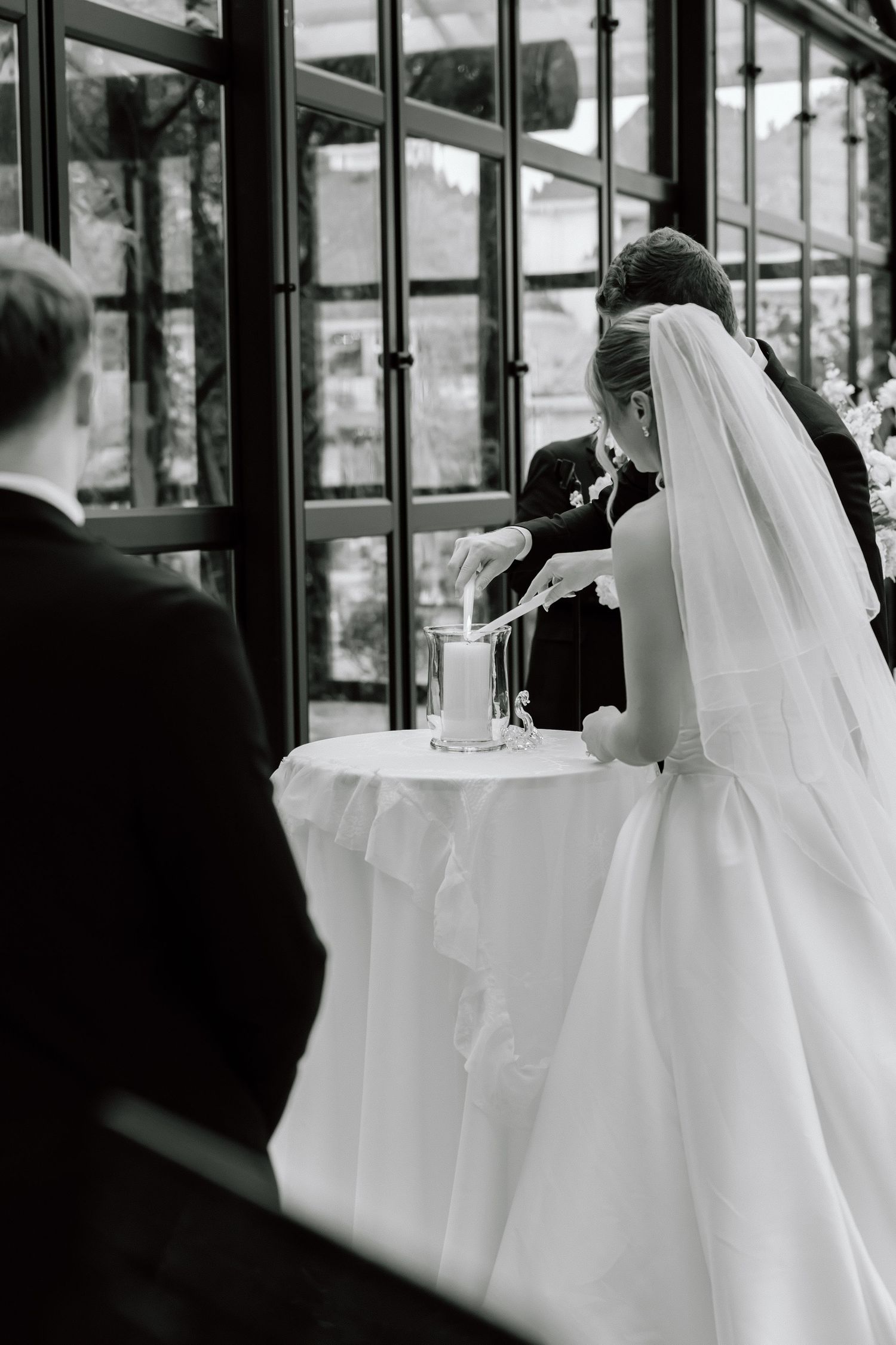 Black and white photo of a bride in a white gown and veil performing a unity candle ceremony at a wedding.