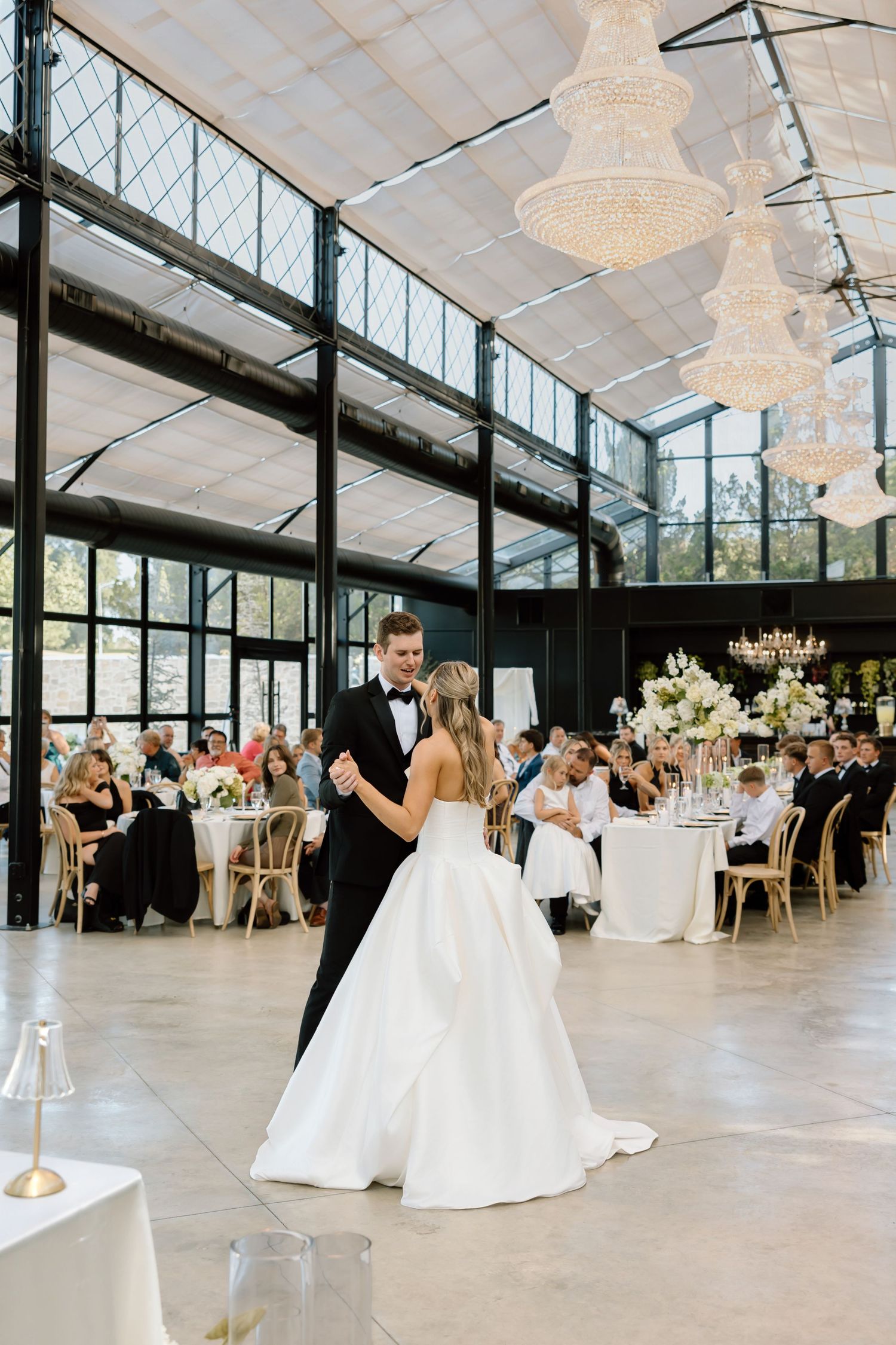 Bride and groom share their first dance in an elegant glass-ceiling venue with crystal chandeliers and seated guests.