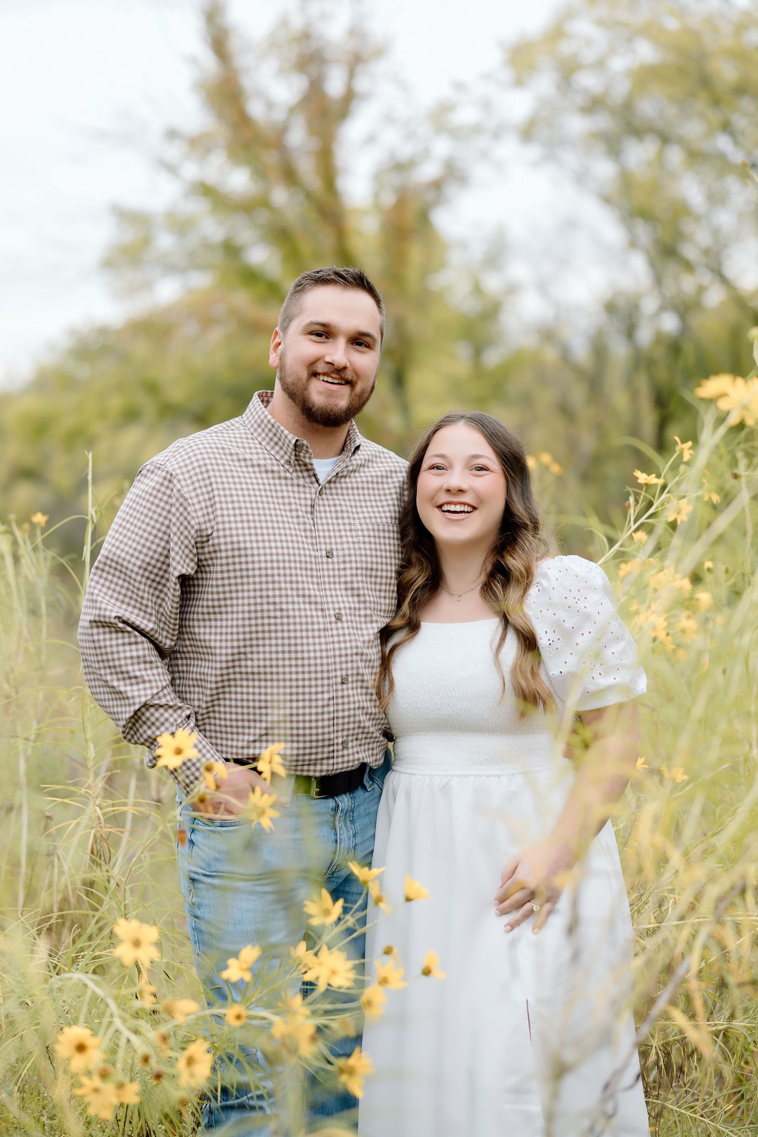A happy couple smiles at the camera while standing in a field of yellow flowers.