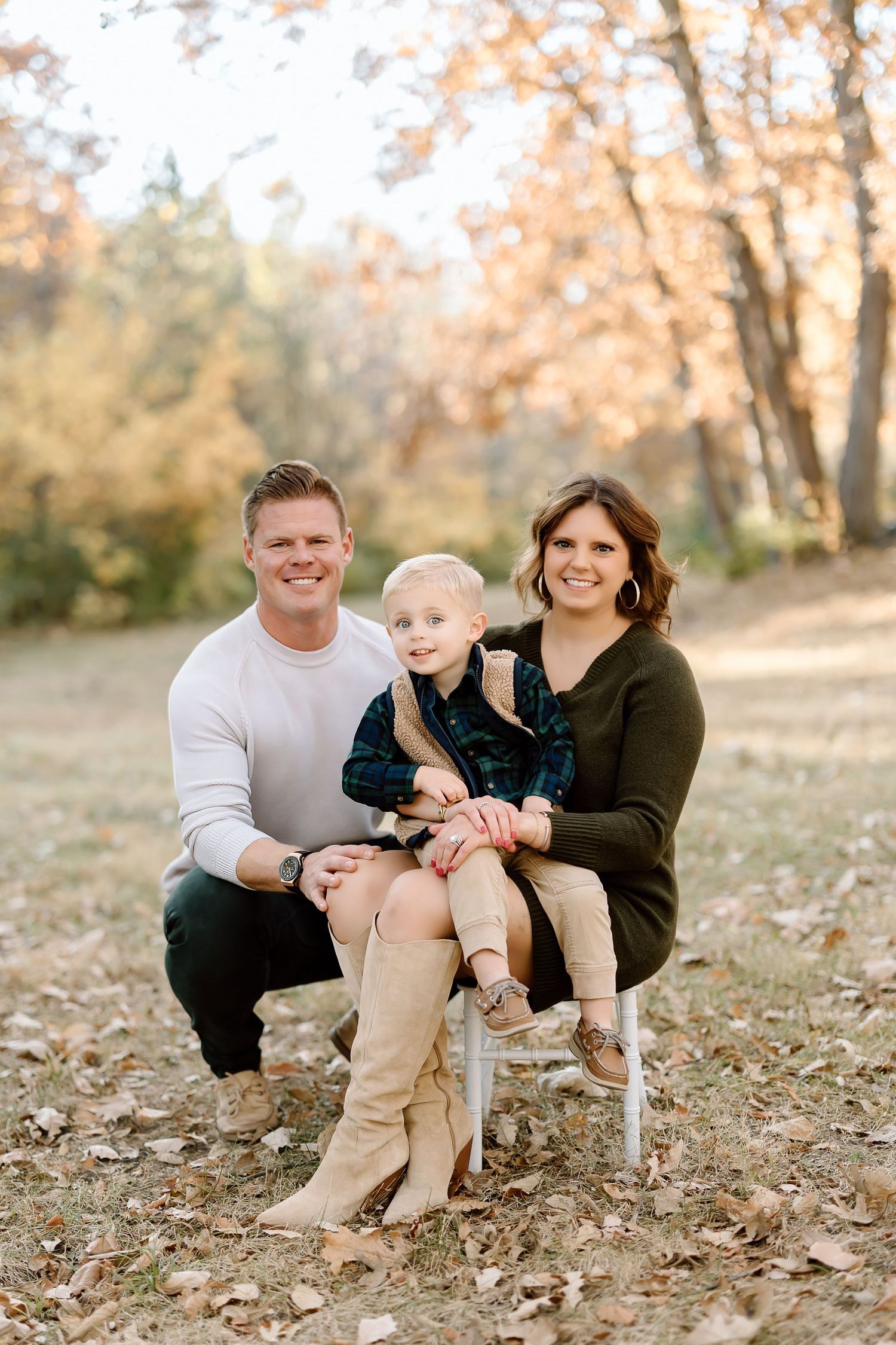 Family of three poses together on white chair in autumn setting.