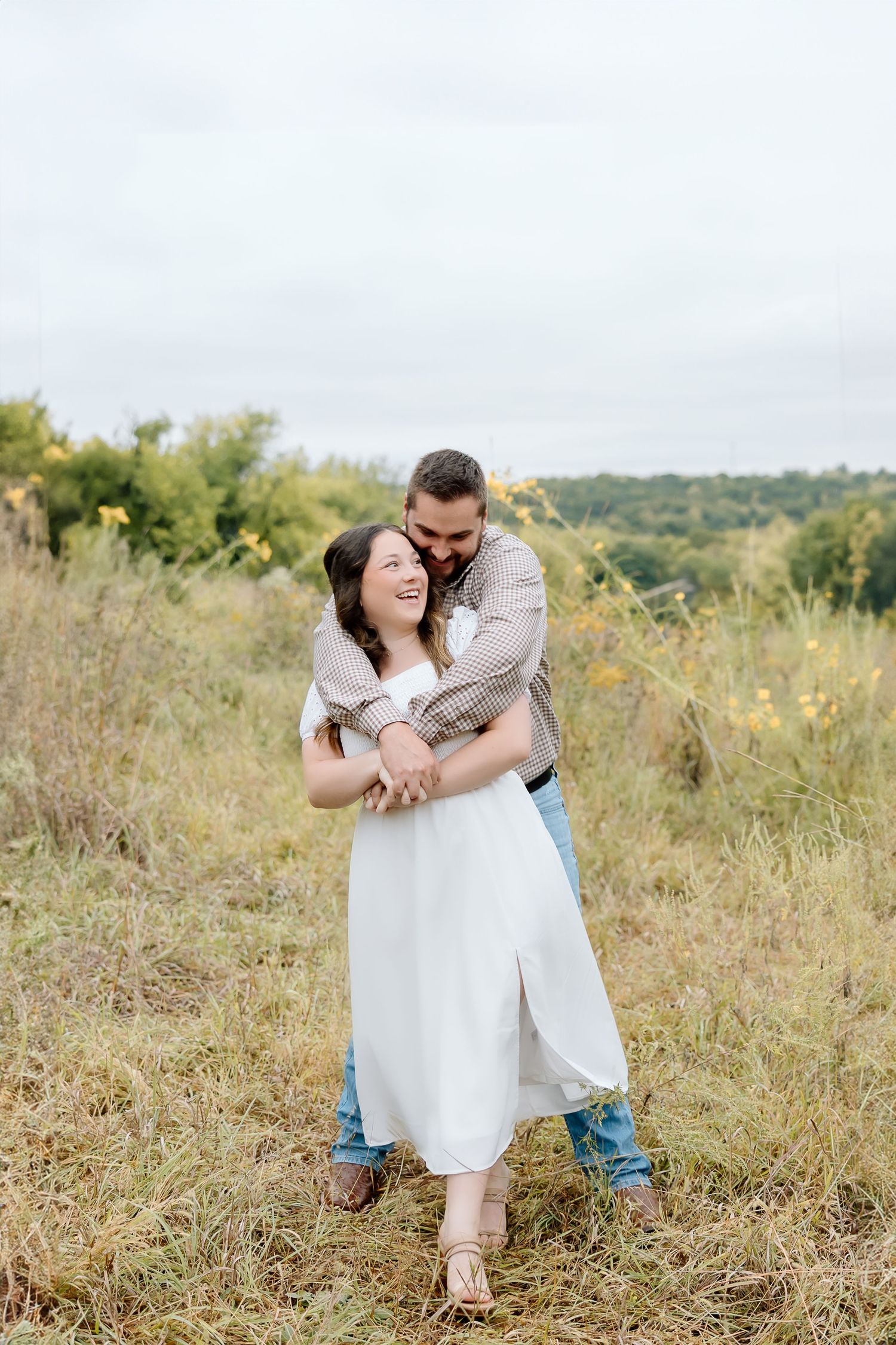 A couple laughs together while embracing in a sunny wildflower field.