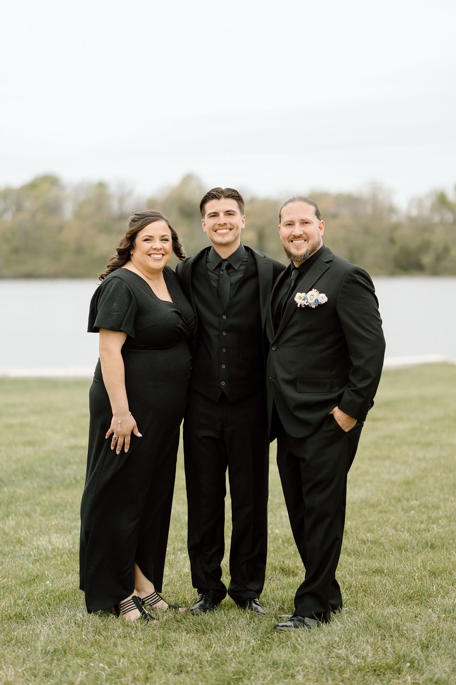 Three people in formal black attire pose together outdoors by a lake.