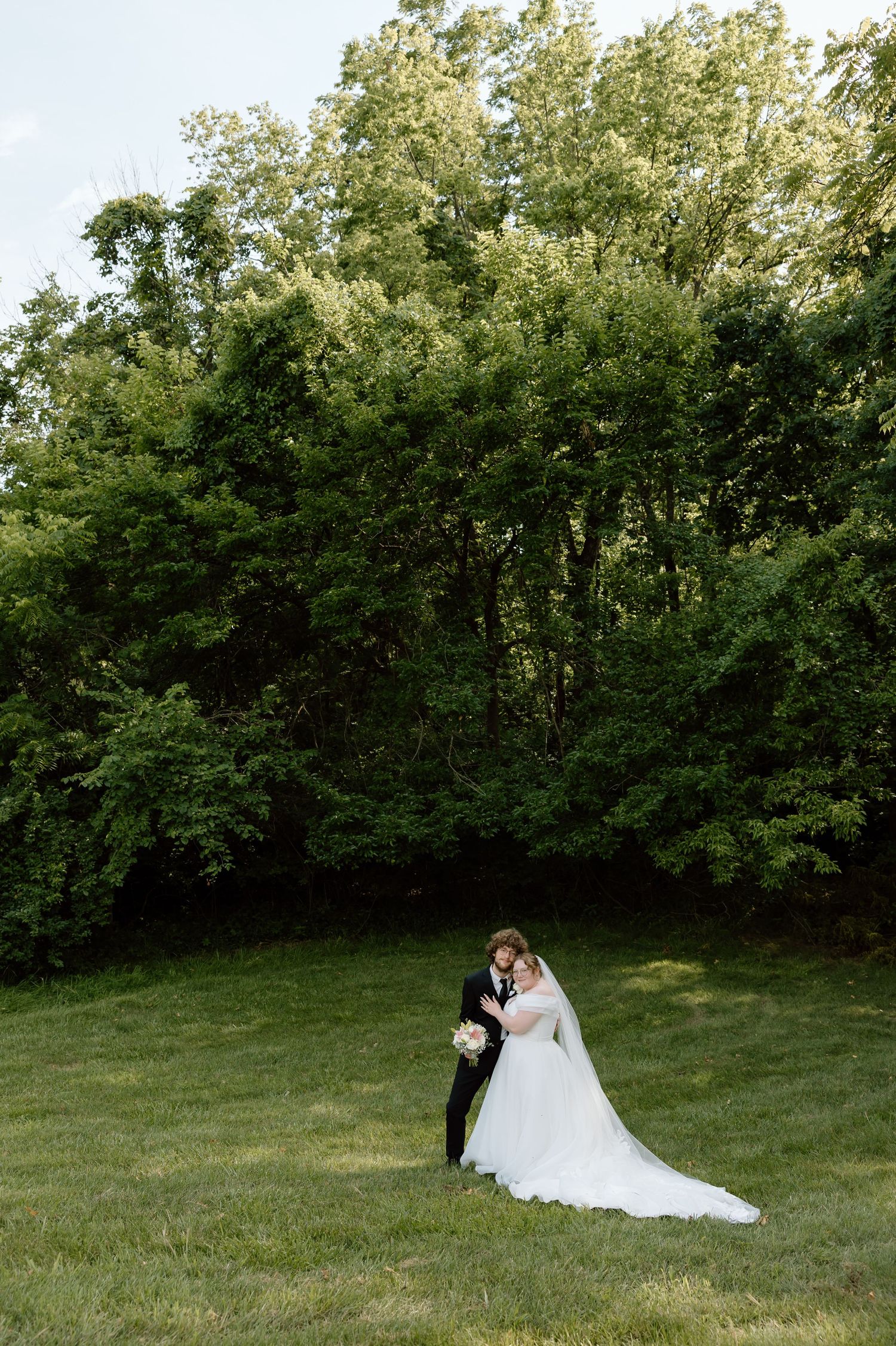 Bride and groom embracing on green lawn with lush trees backdrop on their wedding day.