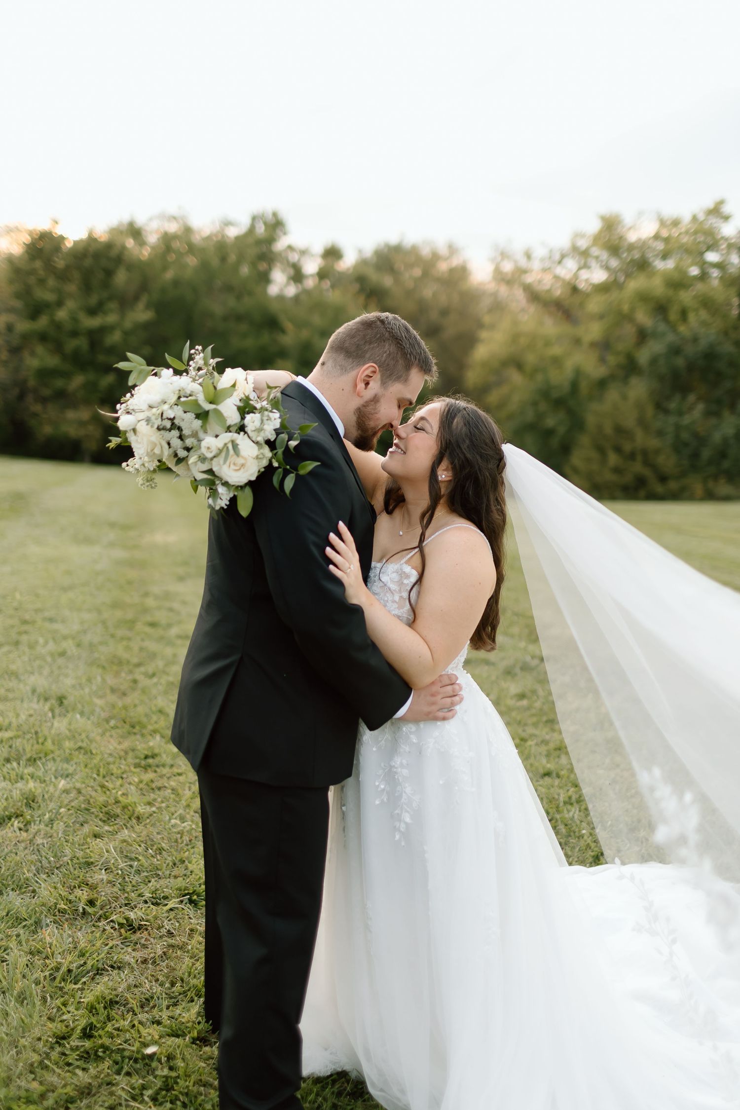 A romantic wedding couple embraces against a sunlit natural backdrop with flowing white veil and floral bouquet.