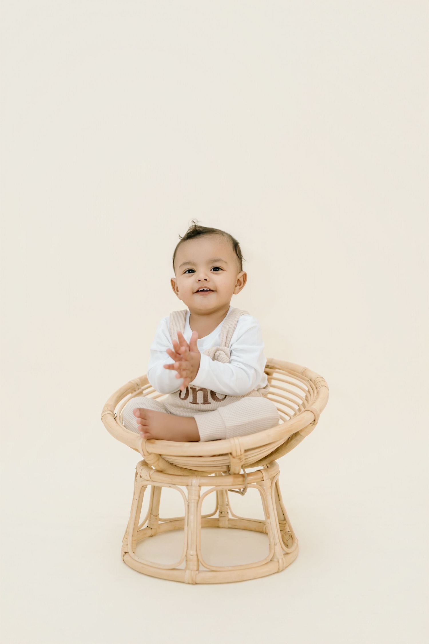 Baby in white outfit sits happily on round rattan chair against neutral background.