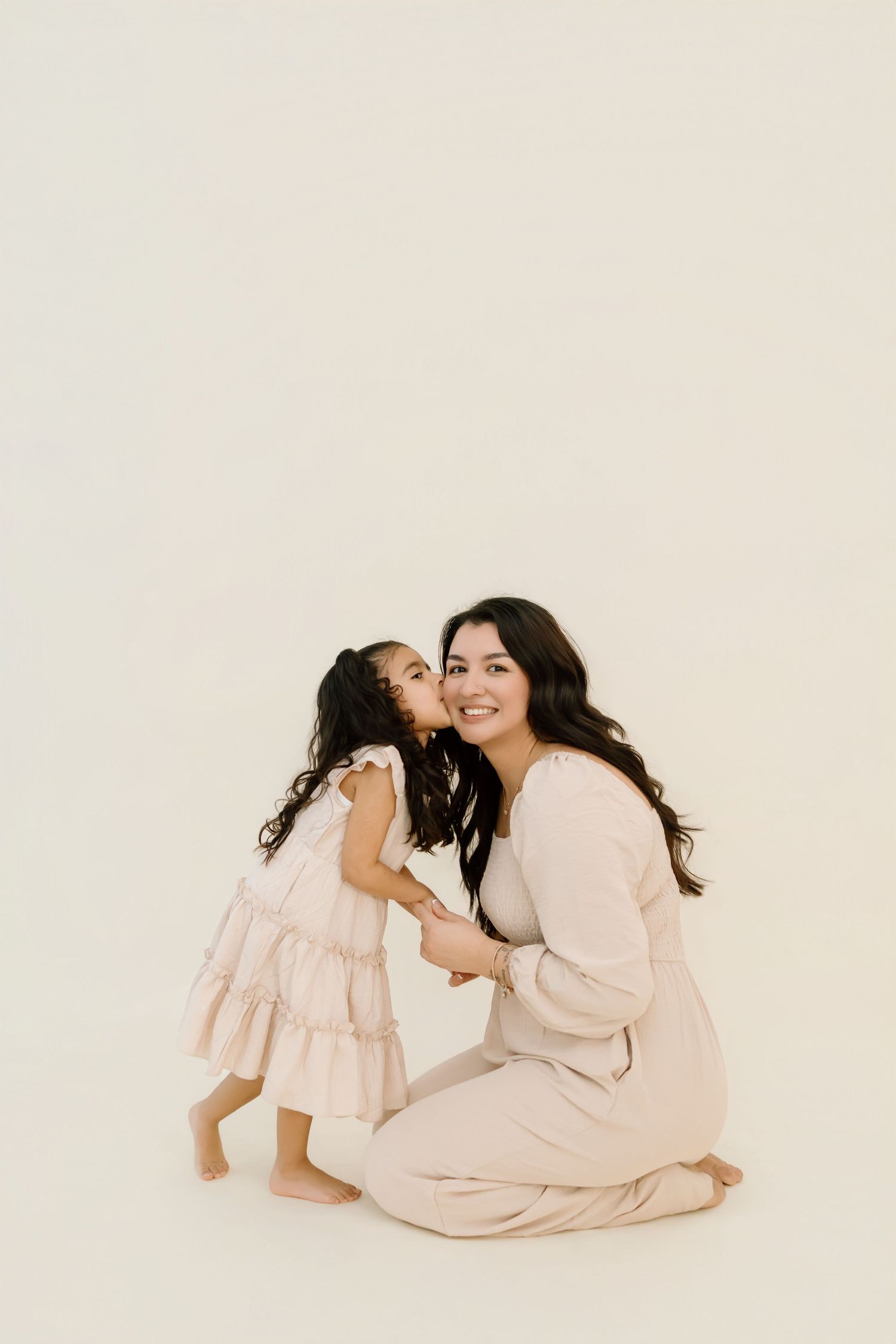 A tender moment between parent and child in matching cream-colored outfits sitting together on a white background.